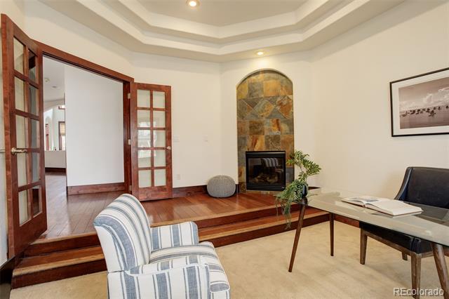 Living room with a stone fireplace, glass-topped desk, striped chair, and raised adjoining room.