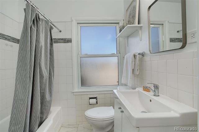 Bathroom with white tile, a gray patterned shower curtain, a wall-mounted sink, and a window.