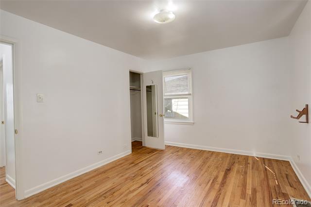 Empty bedroom with polished hardwood floors, an open closet, and a small window with blinds.