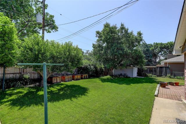 Fenced backyard with a broad lawn, mature trees, garden beds, and a brick patio.