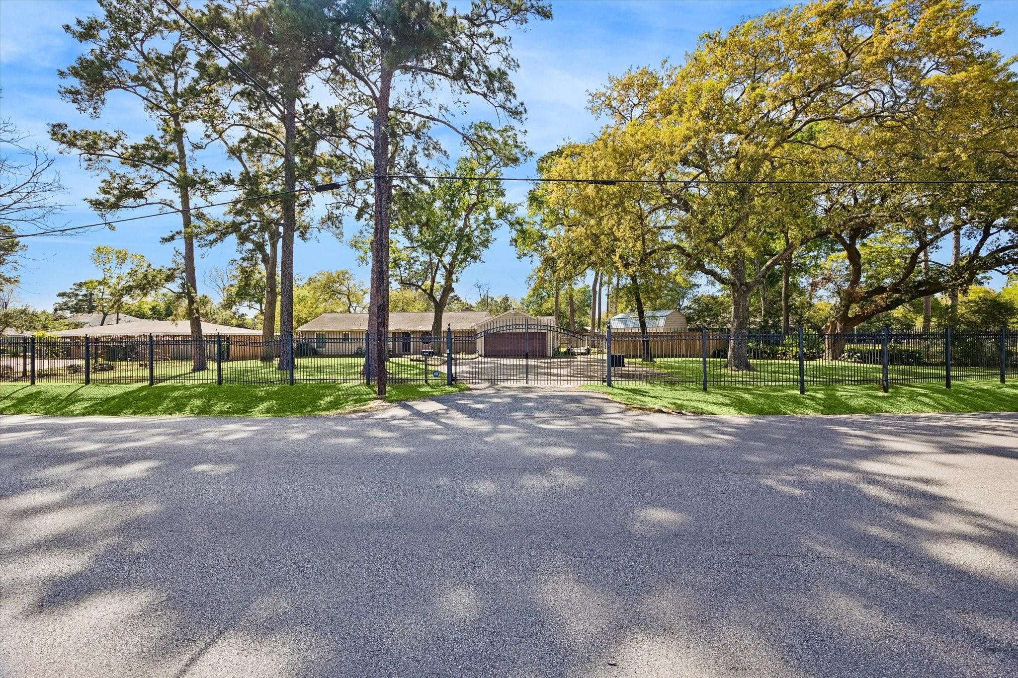 Single-story home behind a black metal fence and driveway gate, shaded by mature trees.