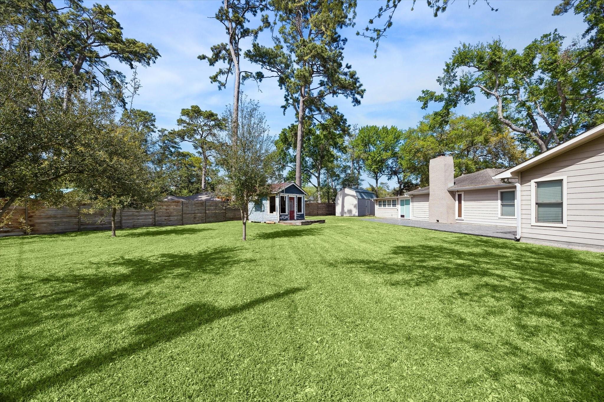 Fenced backyard with a broad lawn, mature trees, small playhouse, and storage shed.