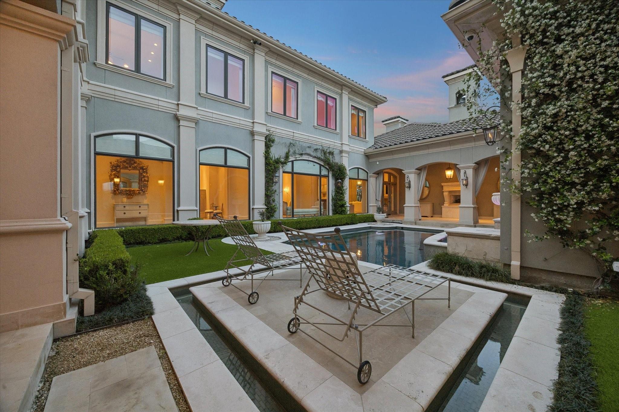 Courtyard pool beside an elegant two-story home, with stone lounge deck and warm window lights at dusk.