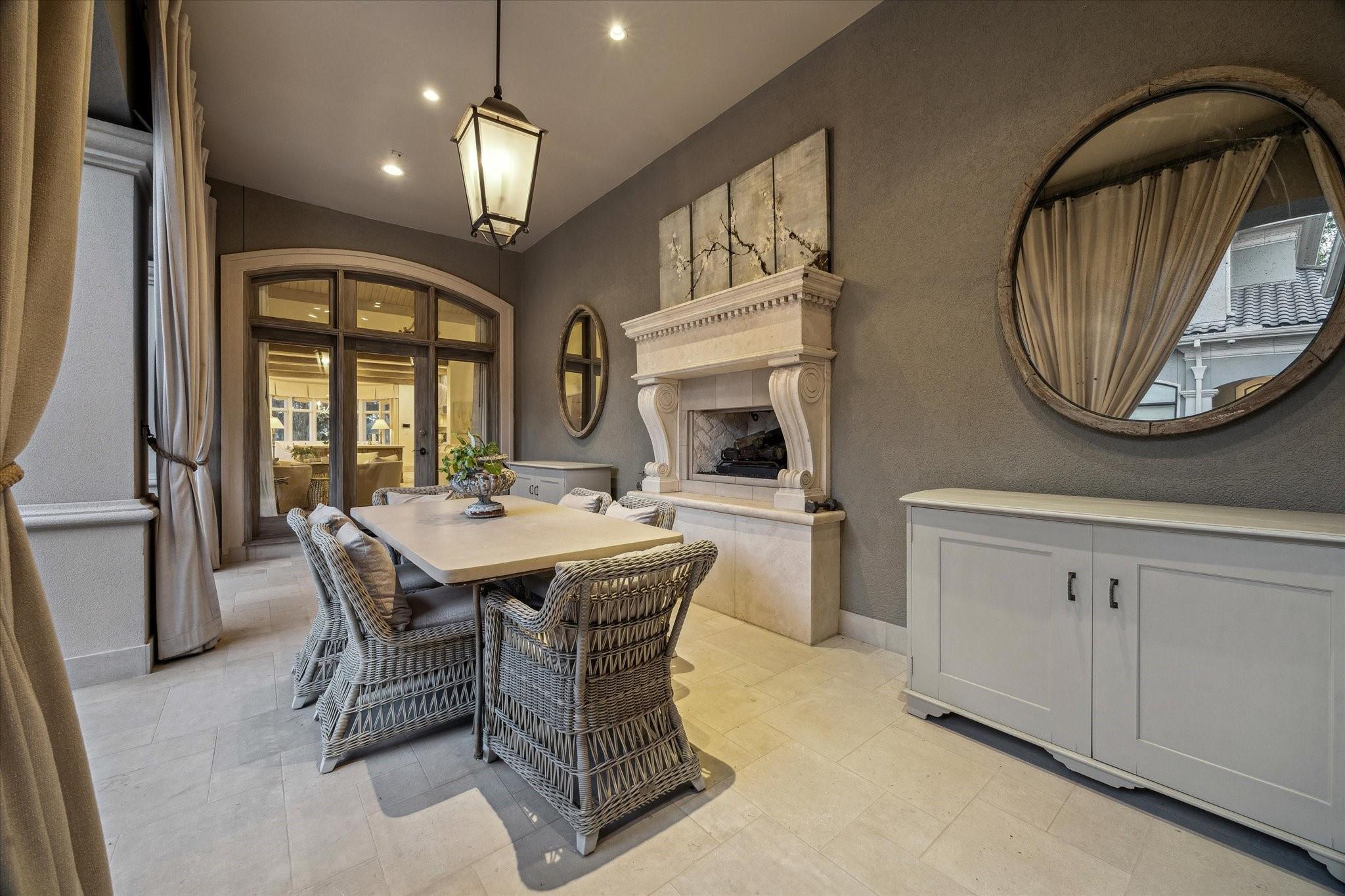 Dining room with woven chairs, an ornate fireplace, arched glass doors, and a large round mirror.