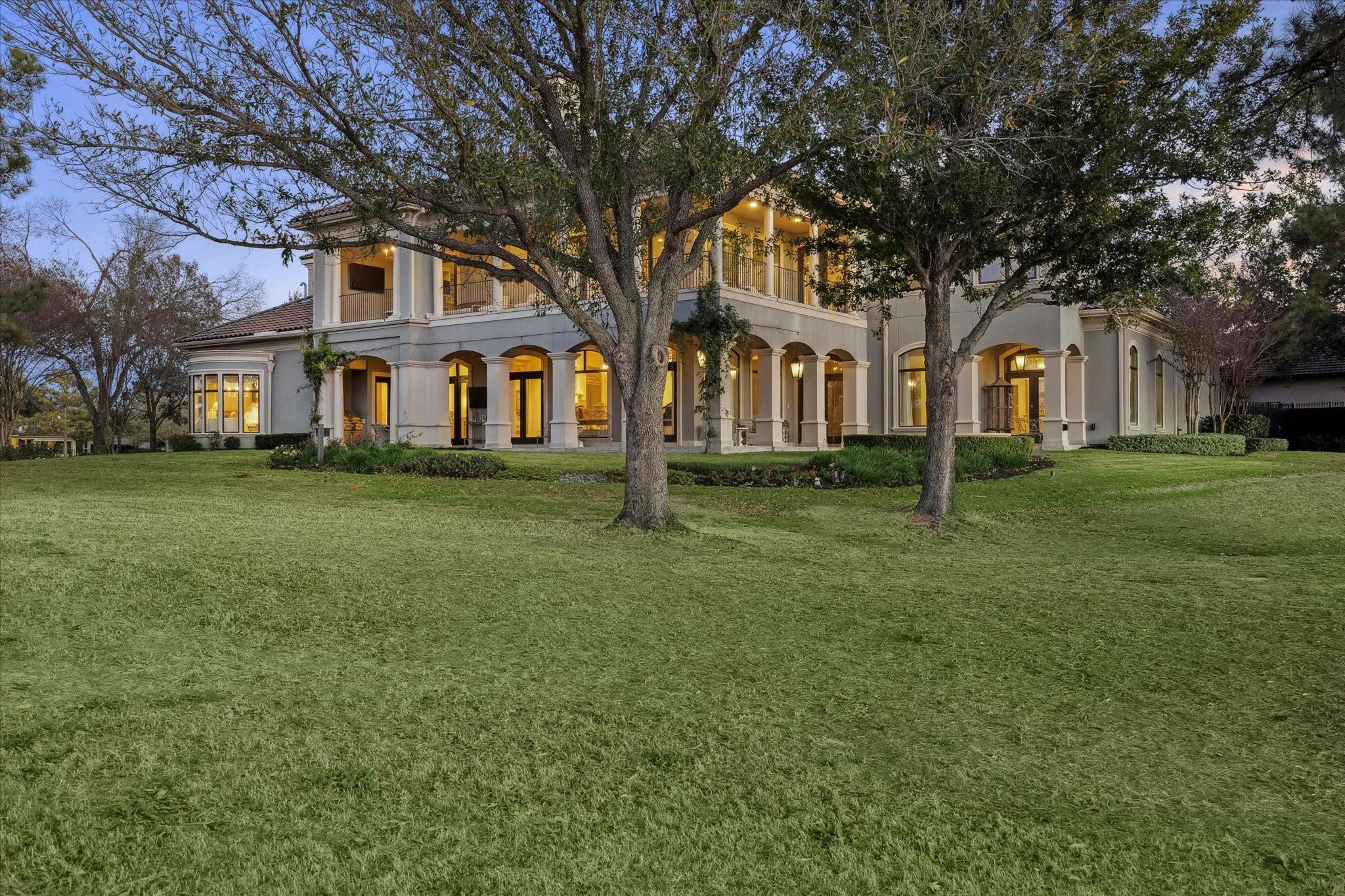 Two-story stucco home with an arched columned patio, mature trees, and a broad lawn at dusk.