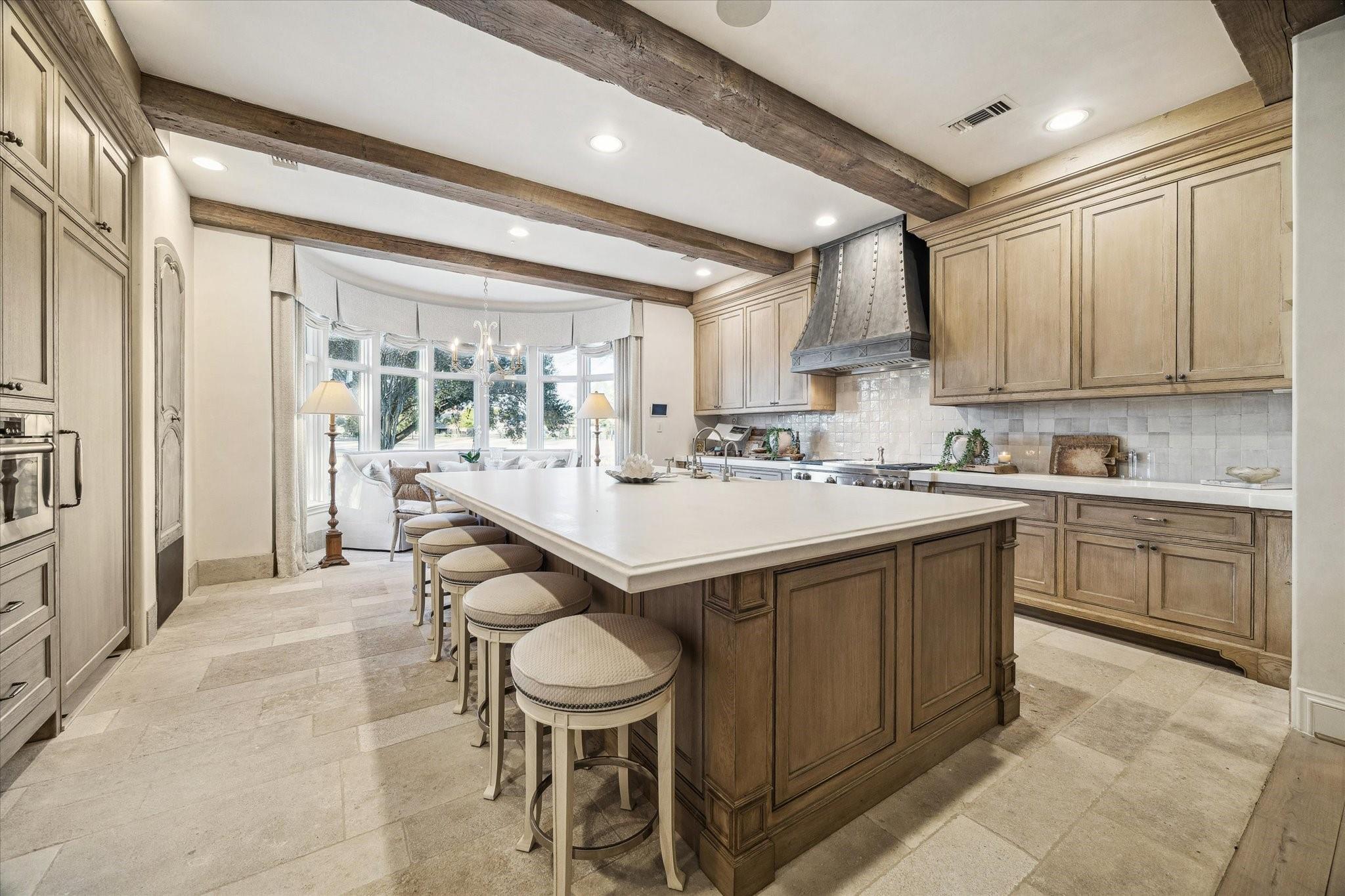 Kitchen with a large wood island and stools beneath exposed beams, beside a bright bay window.