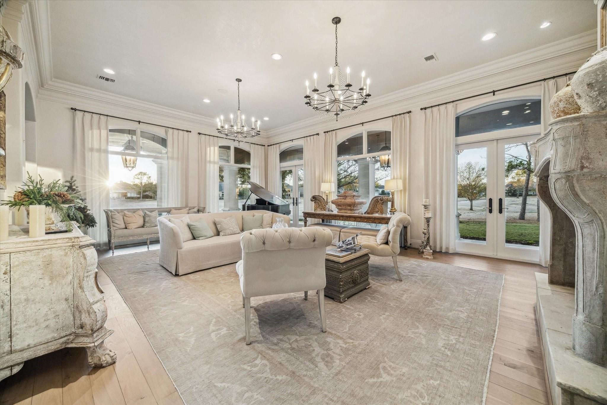Living room with cream seating, two chandeliers, a grand piano, and tall arched windows.