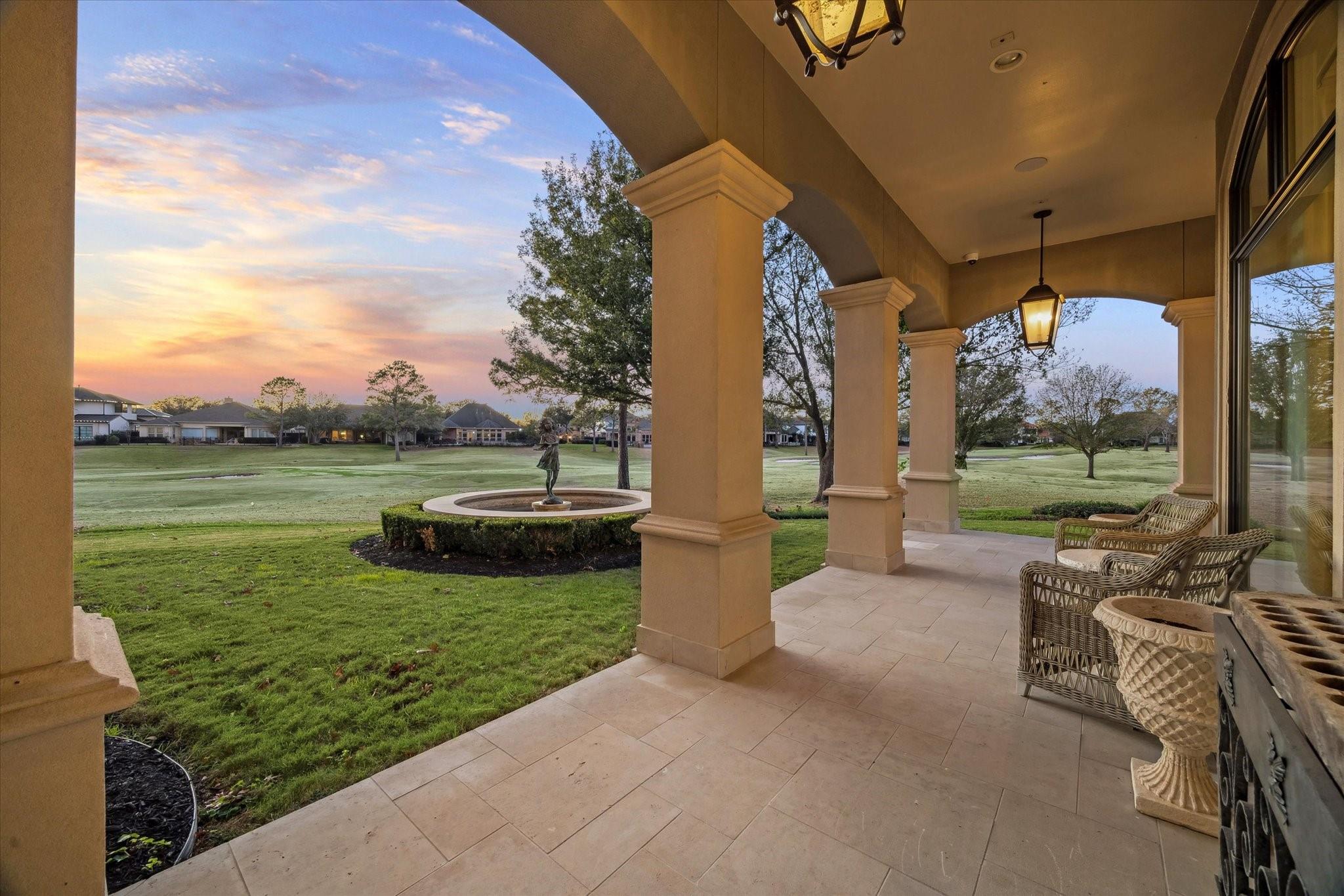 Covered patio with wicker chairs and arched columns overlooking a manicured lawn and circular fountain at sunset.