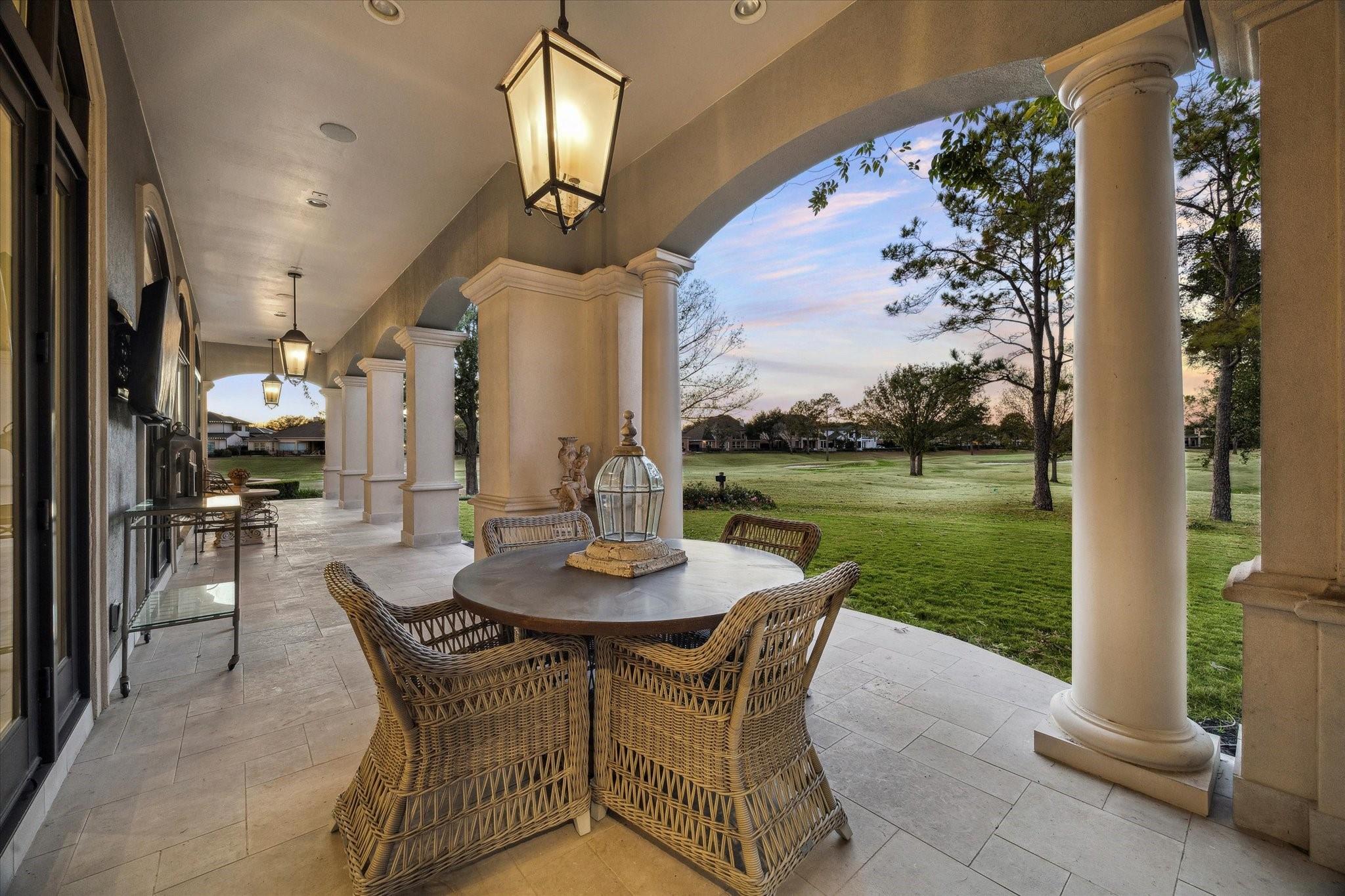 Covered patio with wicker dining set and cream columns overlooking a green lawn at dusk.