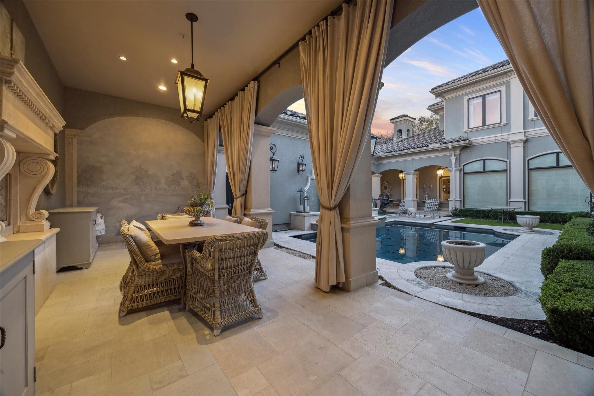 Covered patio with wicker dining set and draped arches overlooking a courtyard pool at dusk.