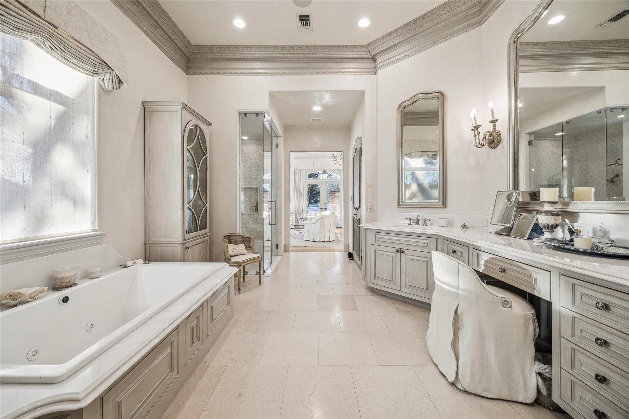Primary bathroom with a built-in soaking tub, long vanity, upholstered chair, and glass-enclosed shower.