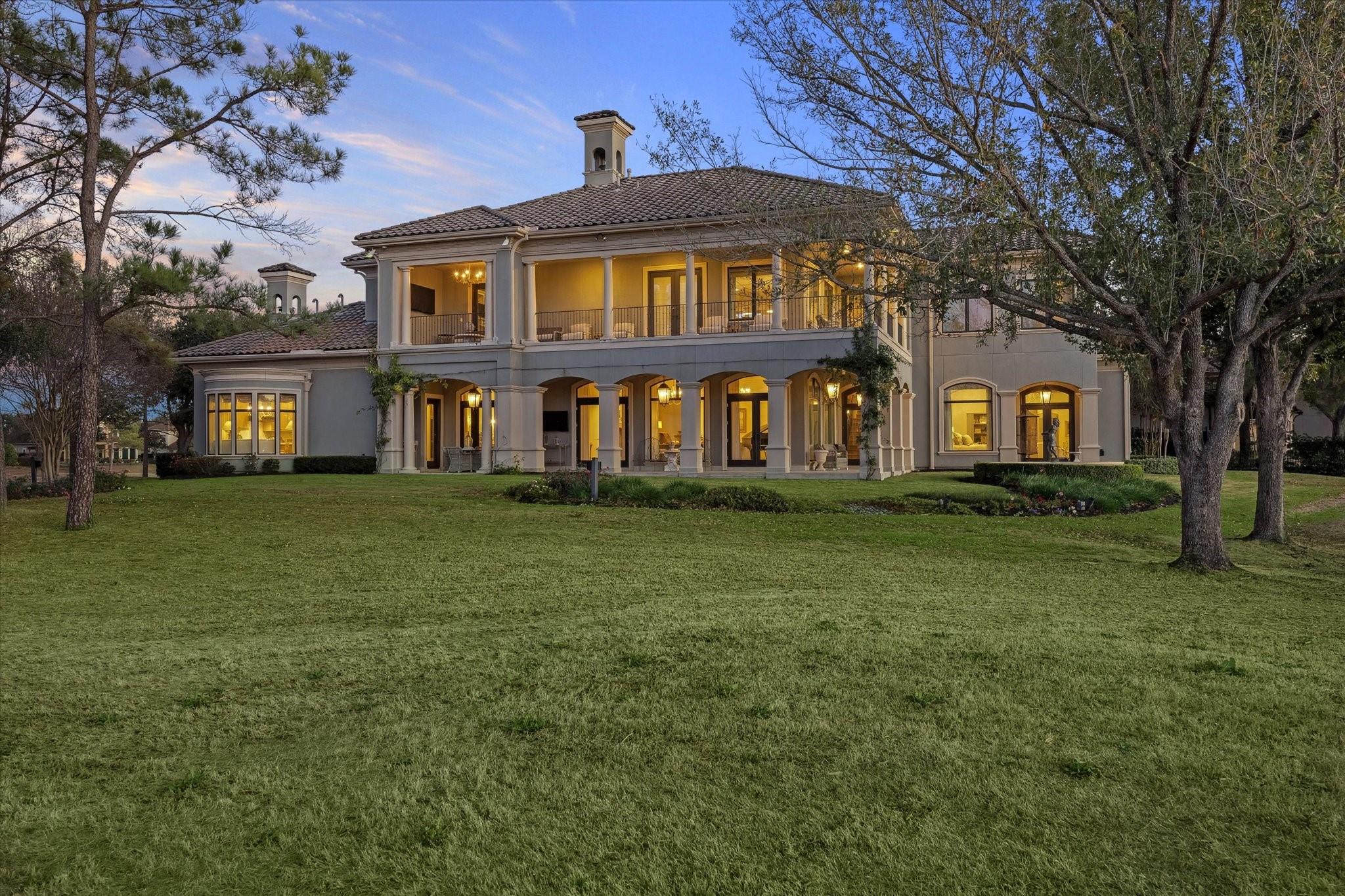 Two-story home with a columned patio and upper balcony overlooking a broad lawn at dusk.