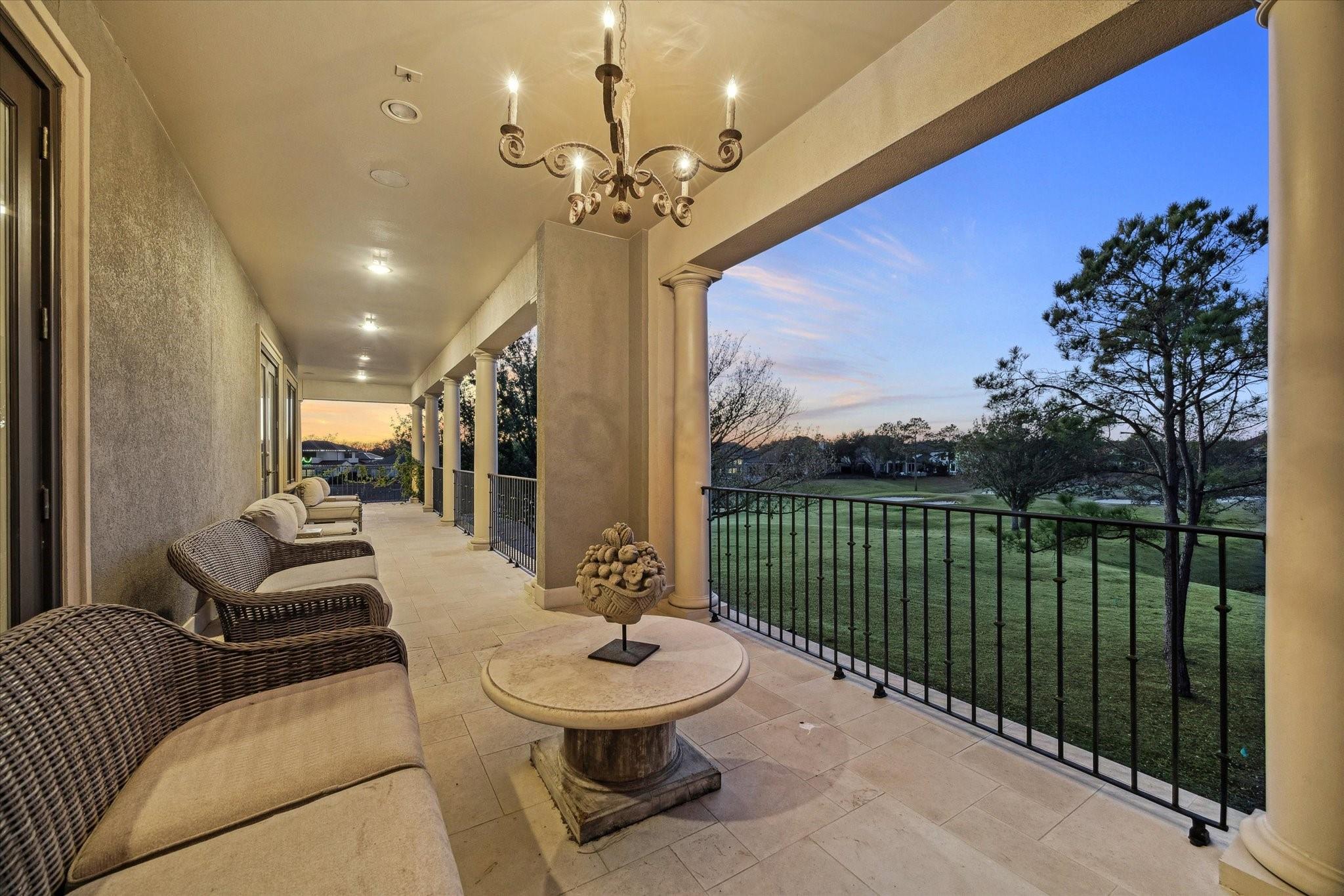 Covered balcony with wicker seating and chandelier overlooking a lawn and water at dusk.