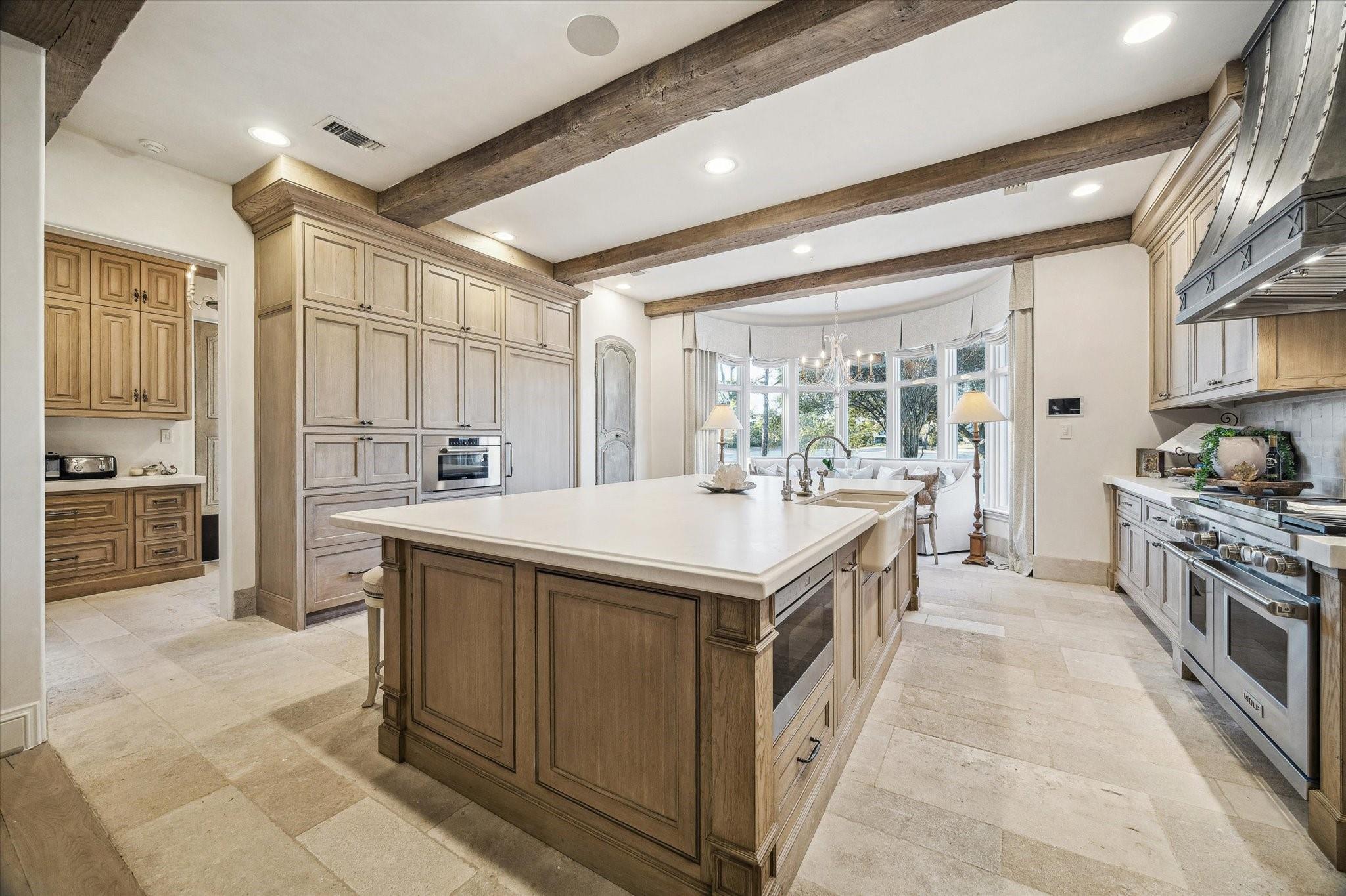 Kitchen with a large wood-paneled island, exposed ceiling beams, and a bright bay-window breakfast nook.