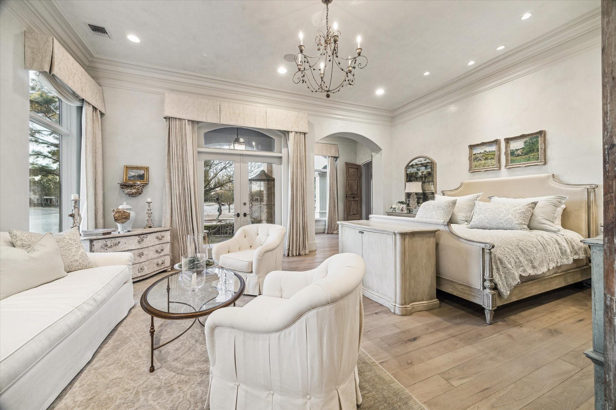 Bedroom with an adjoining sitting area, tall windows, cream furnishings, hardwood floors, and a chandelier.