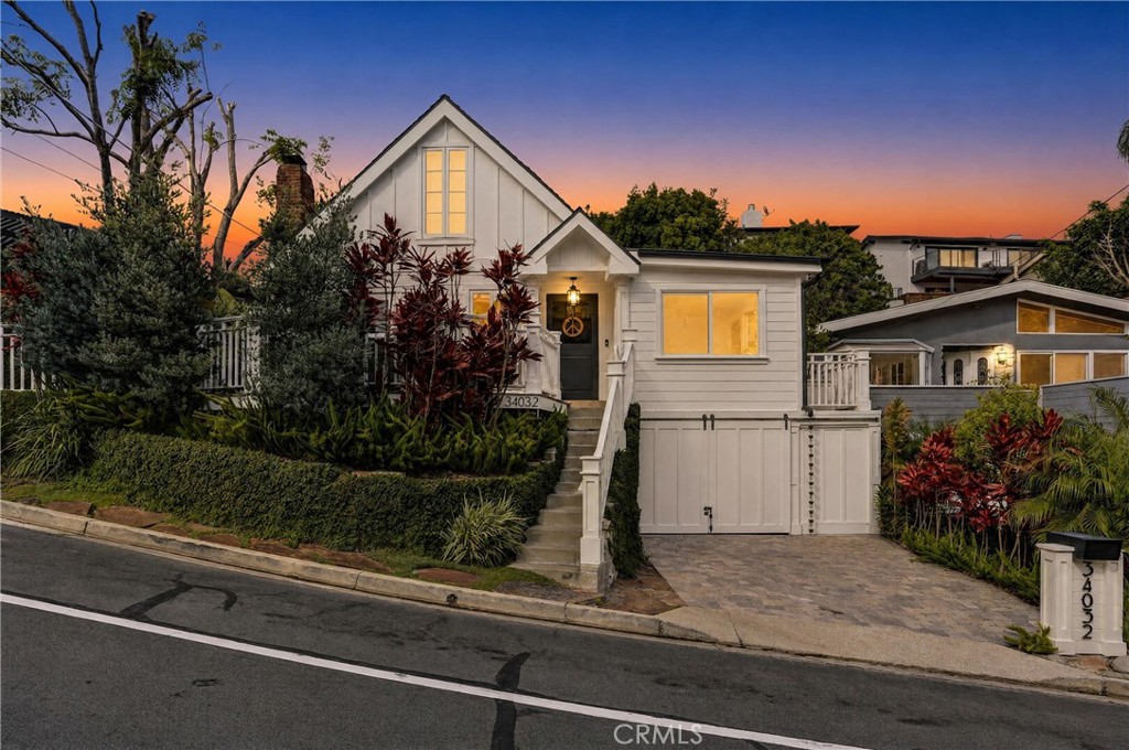 White coastal-style house at dusk, with a steep gabled roof, landscaped entry, and driveway.