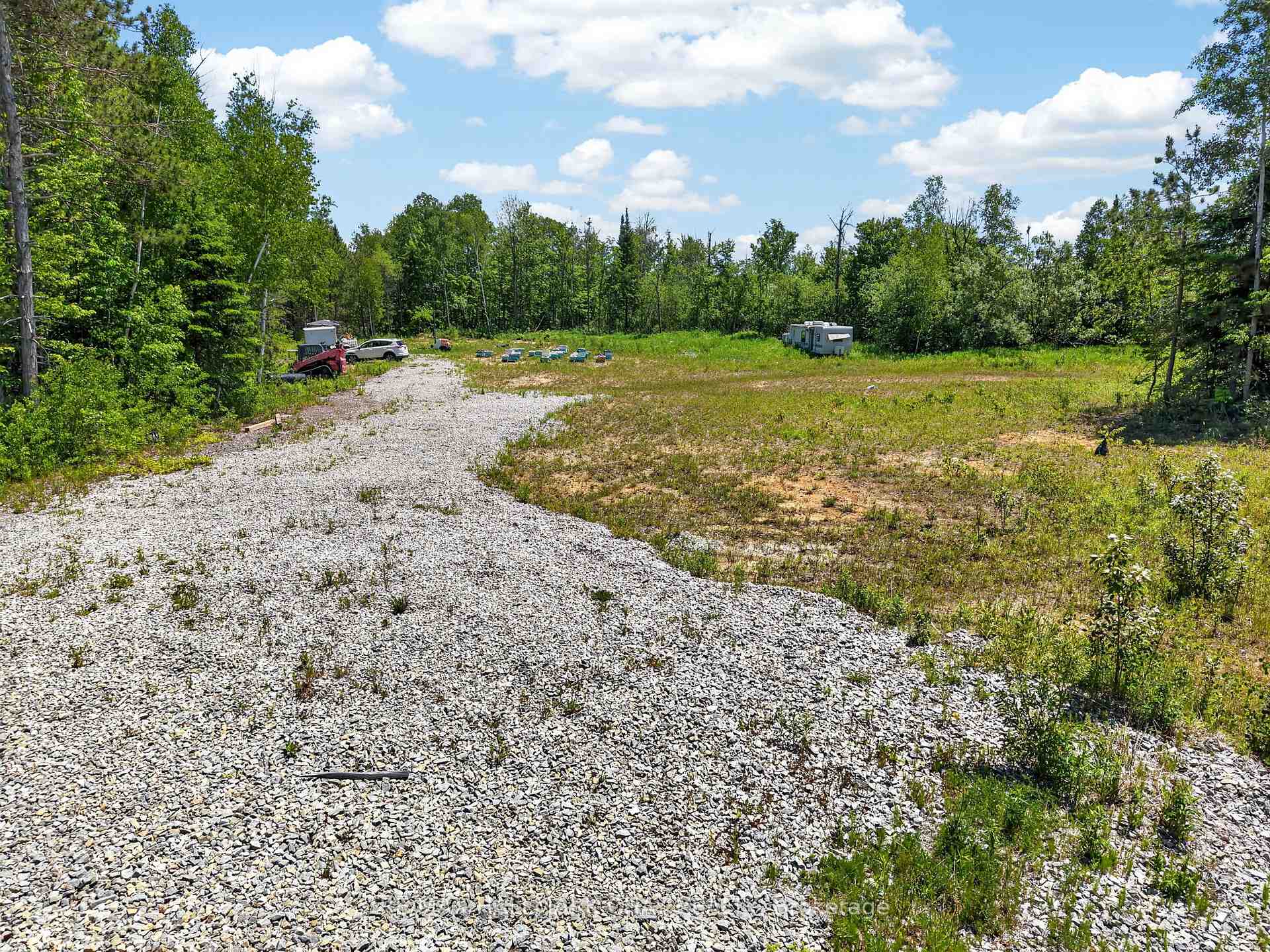 Rural lot with a wide gravel drive, grassy clearing, wooded edges, and parked trailers.