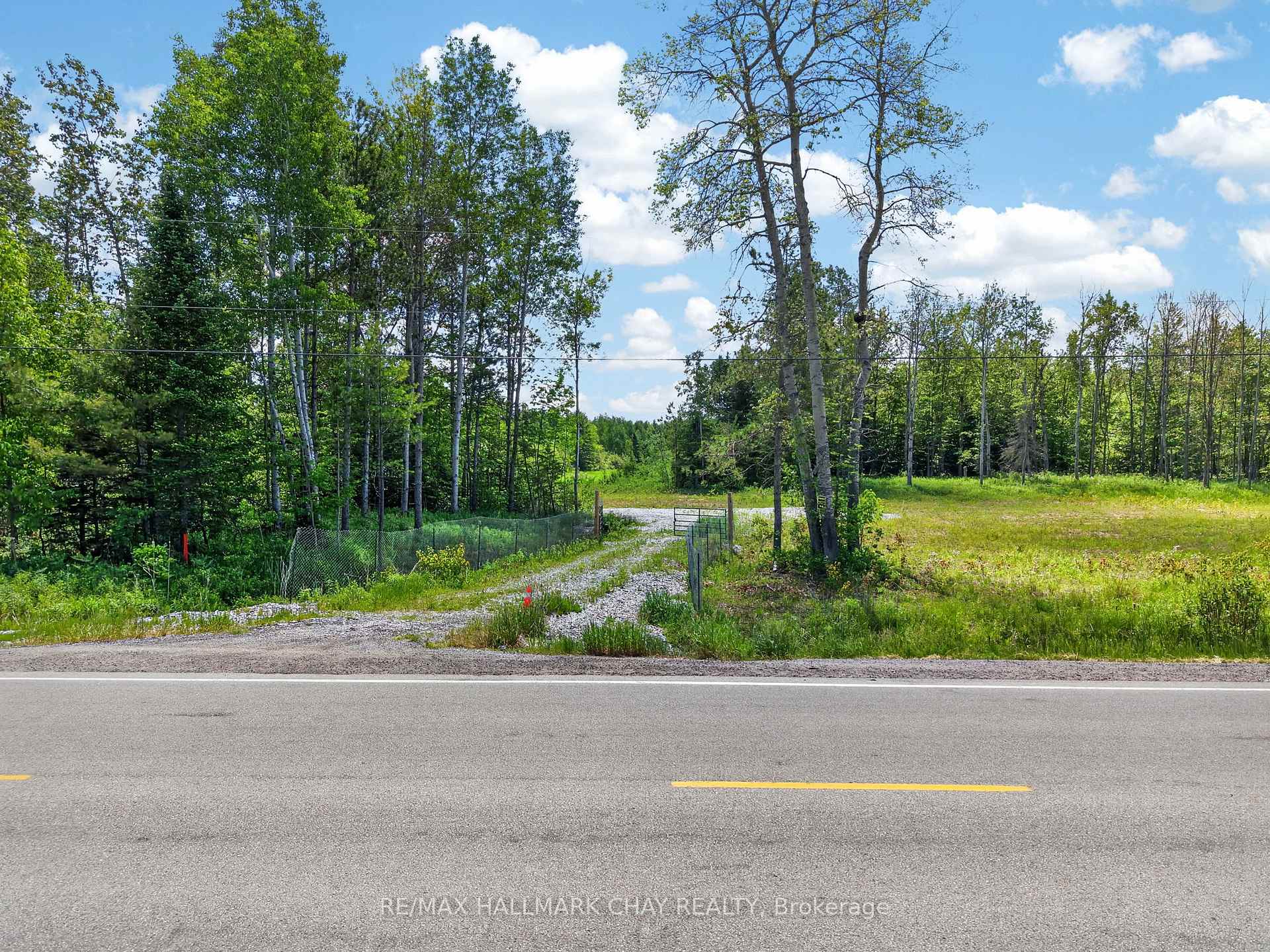 Wooded property entrance with a gated gravel driveway opening onto a paved road.