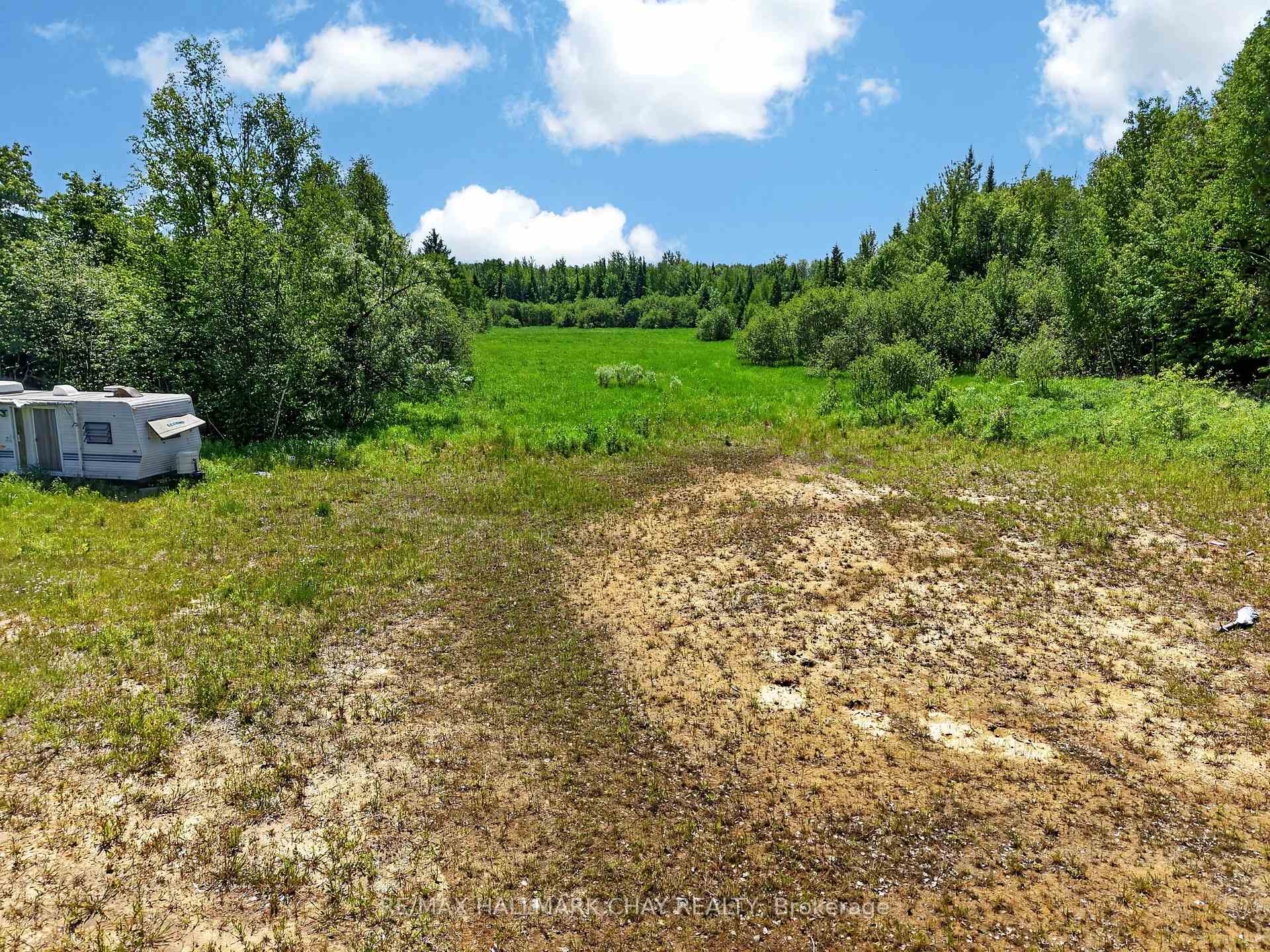 Rural field bordered by trees, with a green meadow and a travel trailer at the edge.