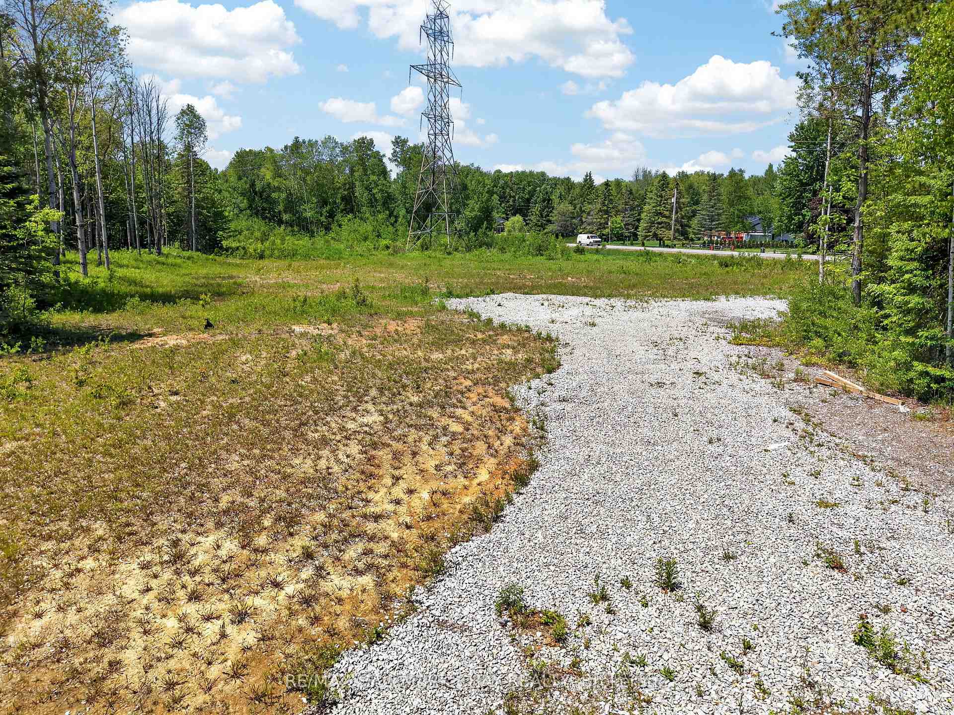 Partly cleared vacant lot with a wide gravel driveway and a utility tower near the road.