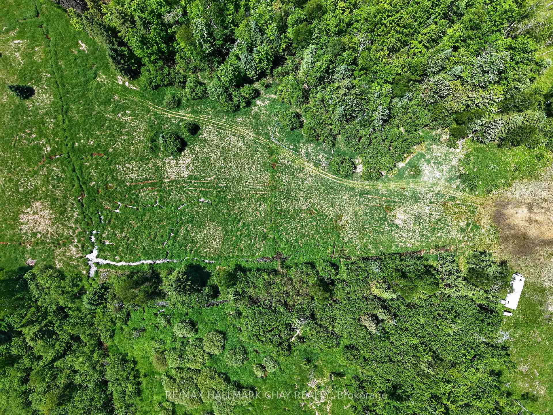 Aerial view of open fields bordered by dense woodland, with a stream and small white structure.