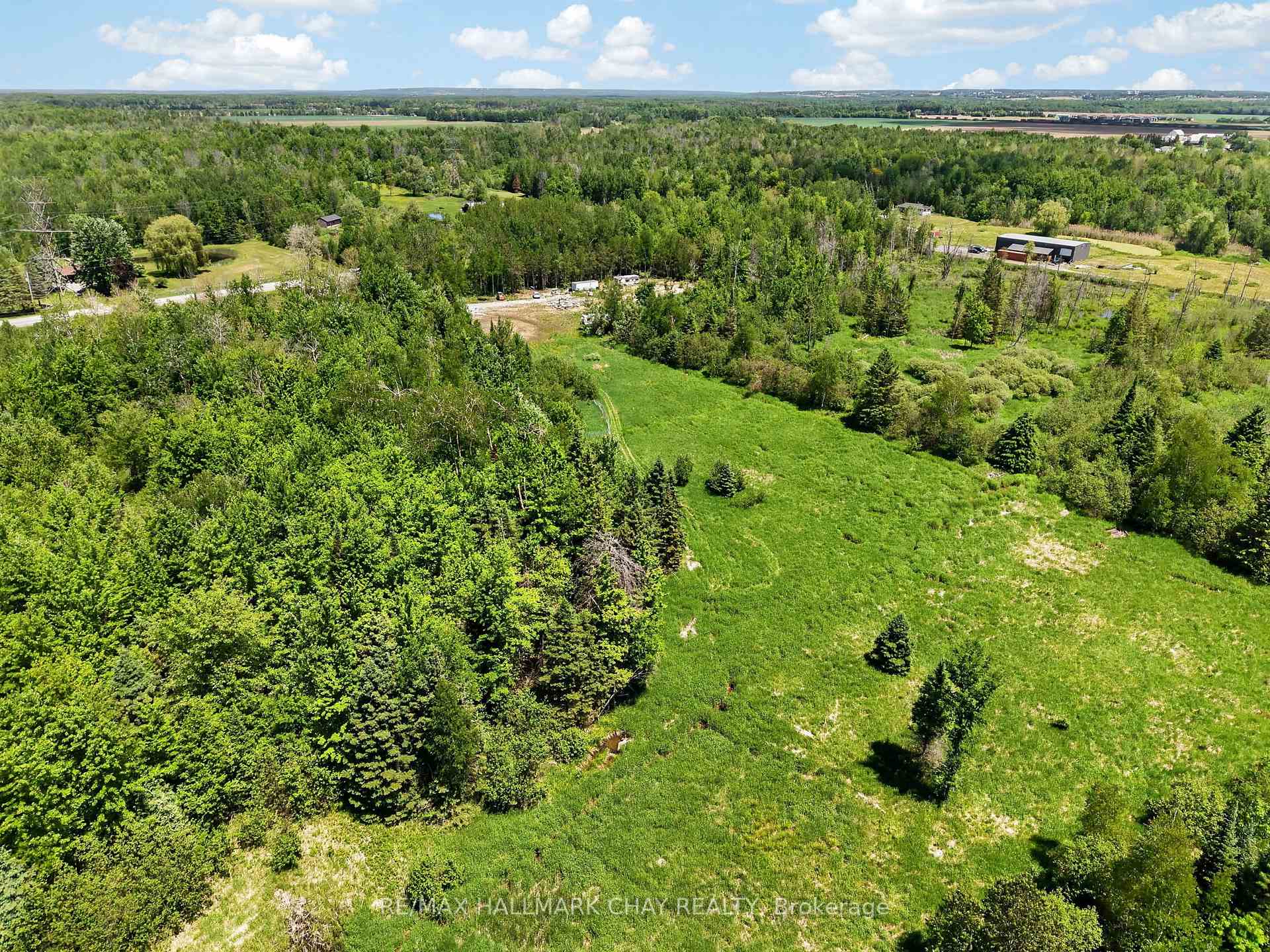 Aerial view of a grassy clearing bordered by dense woodland, with a road and outbuildings in the distance.
