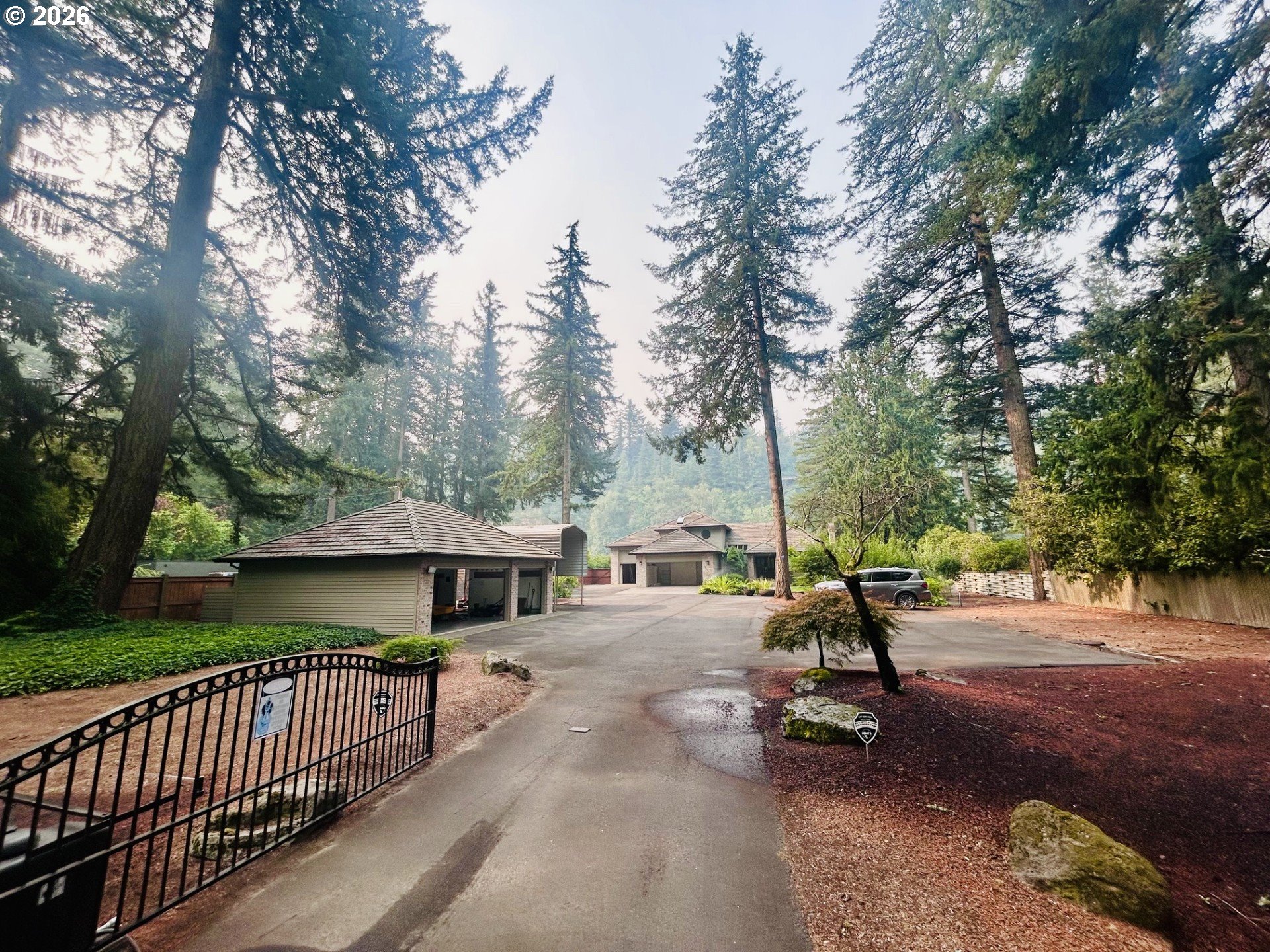 Gated driveway and multiple buildings nestled among tall evergreen trees.