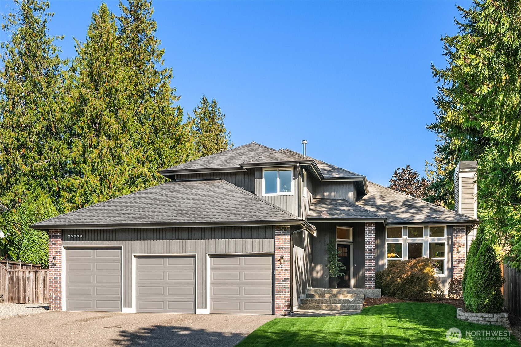 Gray-and-brick two-story home with a three-car garage, landscaped lawn, and tall evergreens.