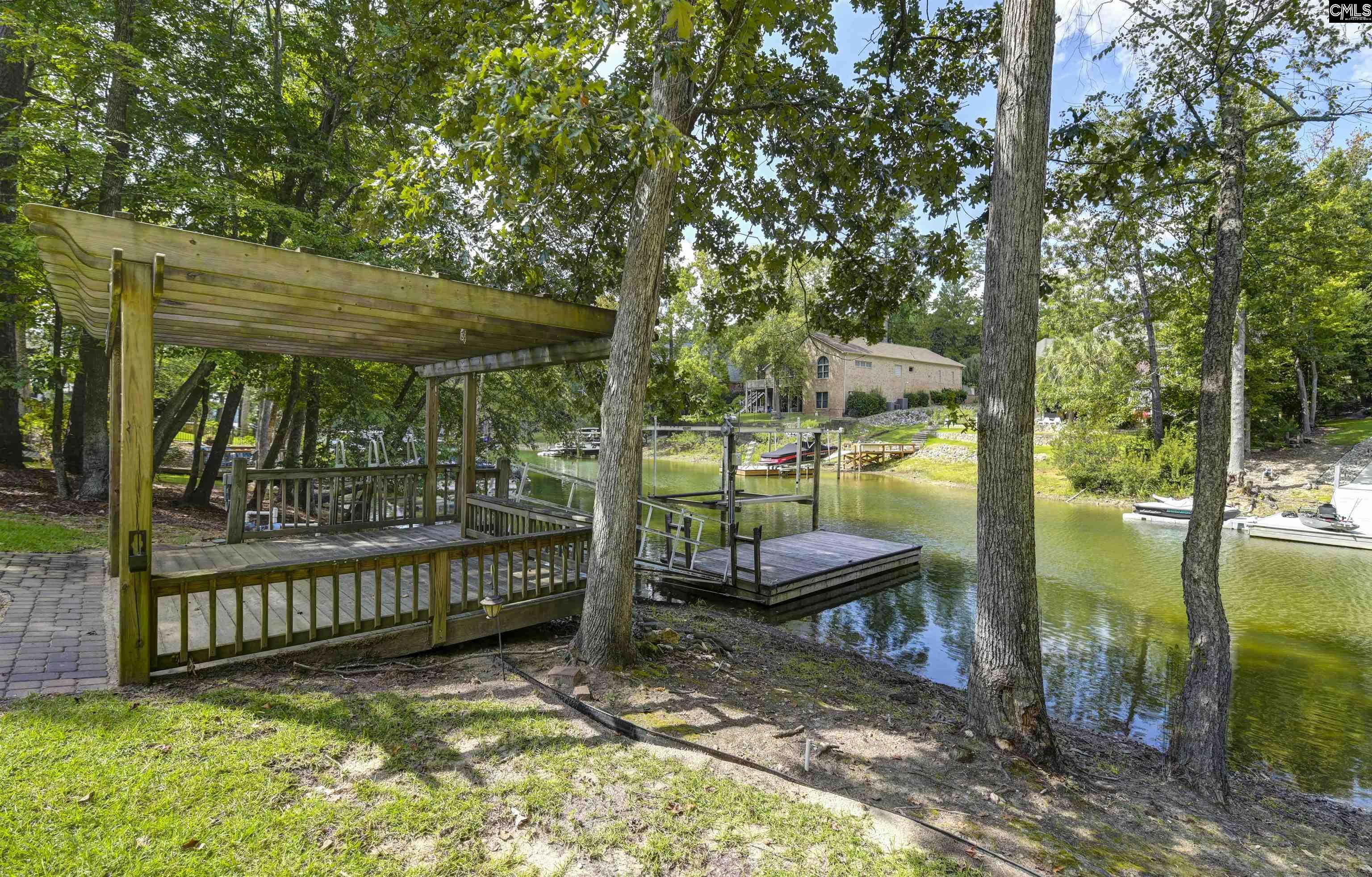 Covered wooden dock beside a tree-lined waterfront, with a floating platform and brick home across the water.