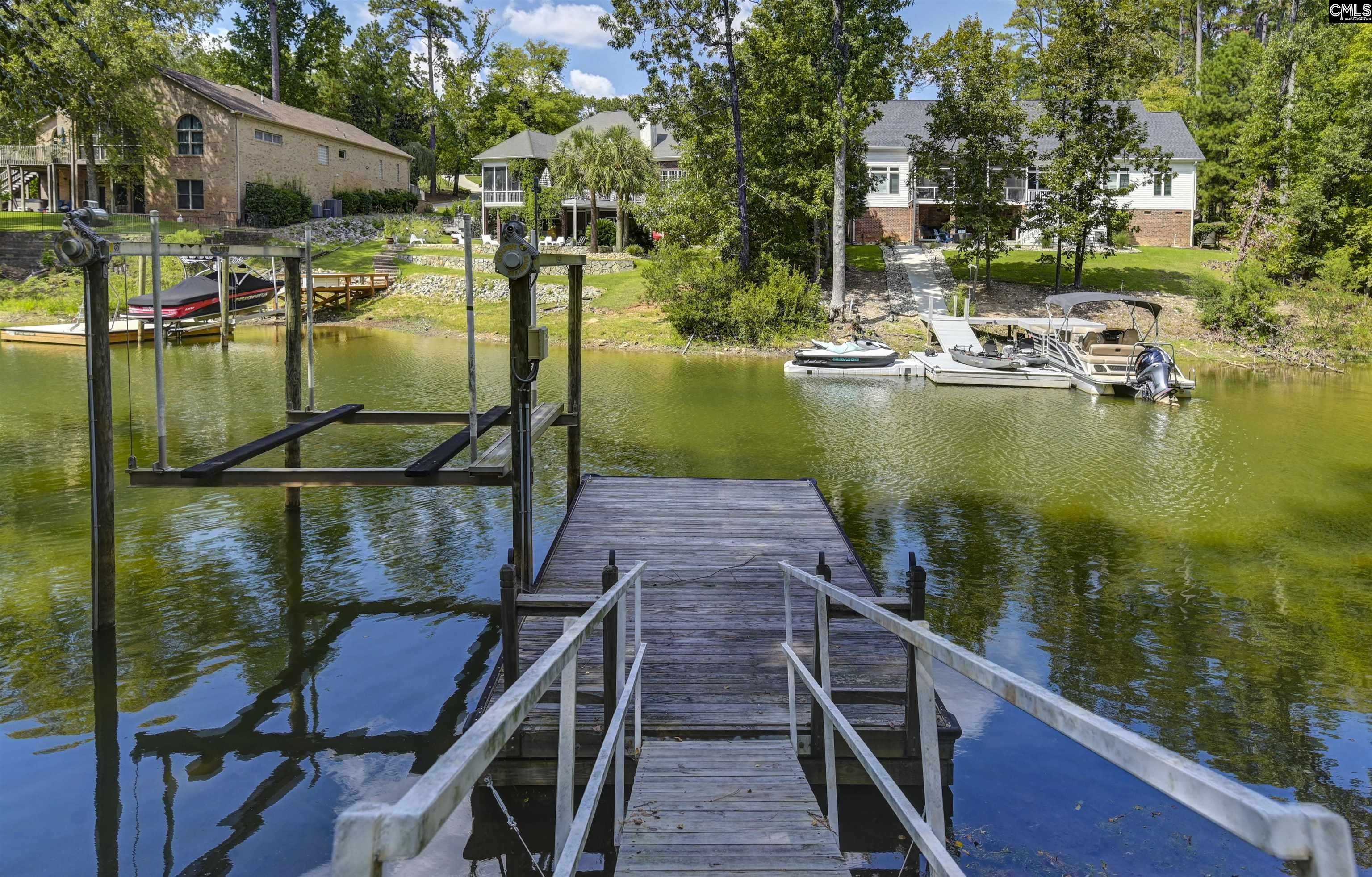 Lakefront dock with a boat lift, boats across the water, and wooded homes beyond.