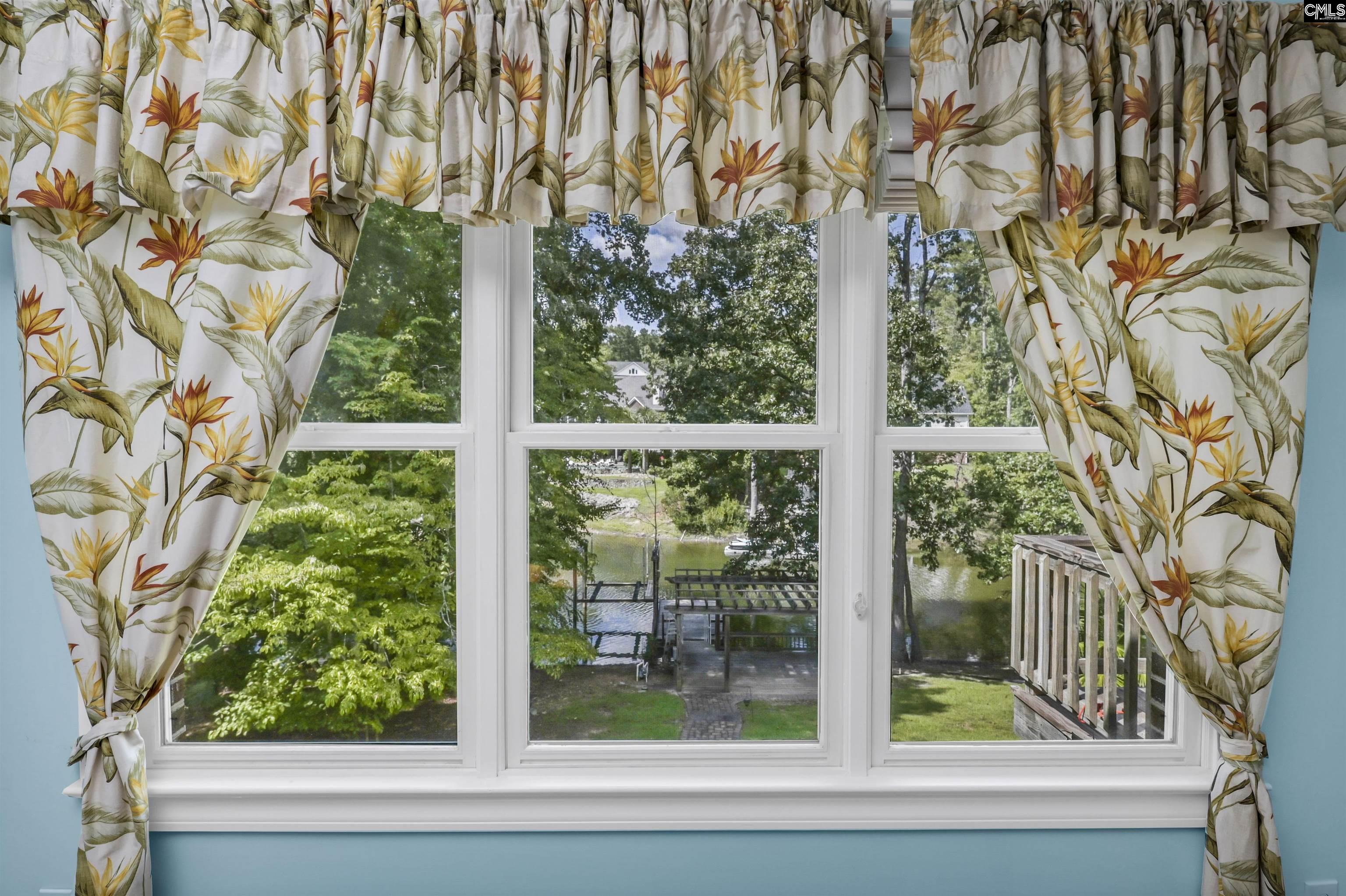 Window with botanical curtains overlooking a leafy lakeside yard, dock, and water.