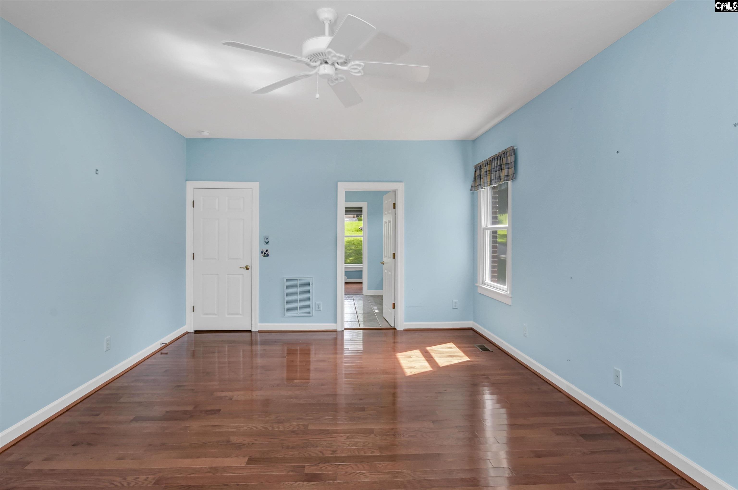 Vacant bedroom with light blue walls, hardwood floors, a ceiling fan, and sunlight through a window.