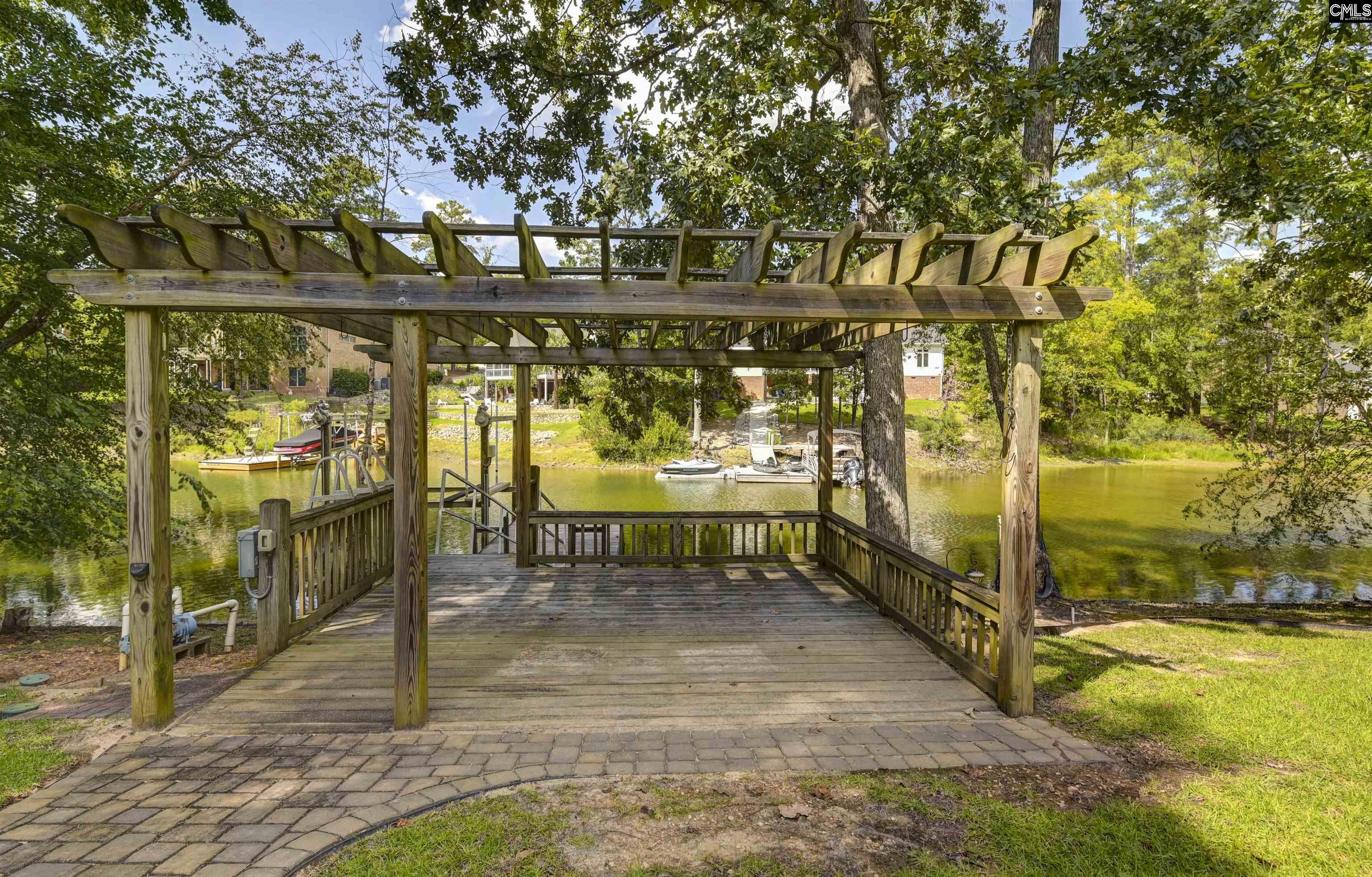 Wooden lakeside dock beneath a pergola, with boats and wooded shoreline across the water.