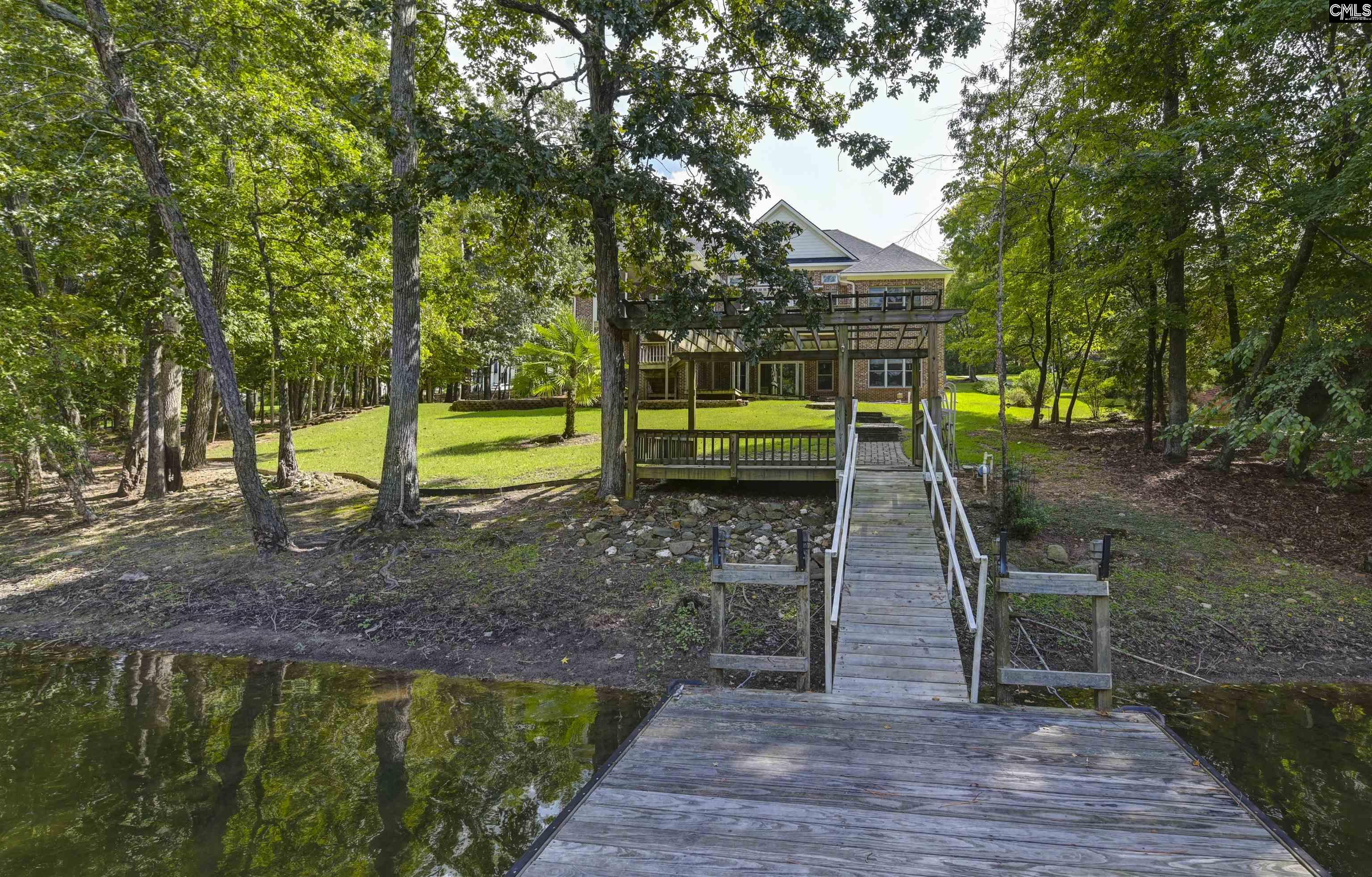 Brick waterfront home beyond a wooden dock and walkway, with a raised deck and wooded lawn.