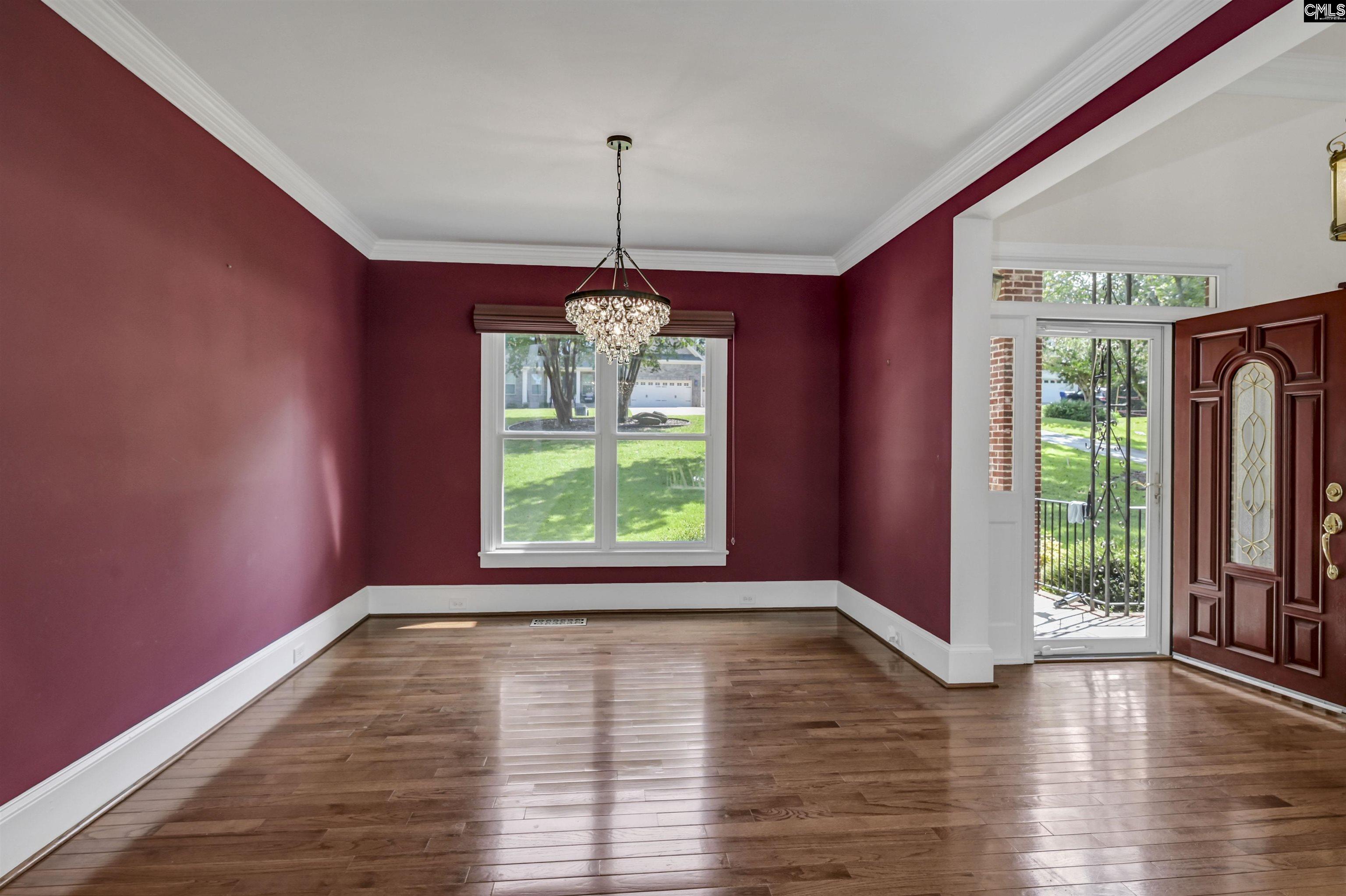 Empty dining room with burgundy walls, hardwood floors, and a crystal chandelier beside the open front door.