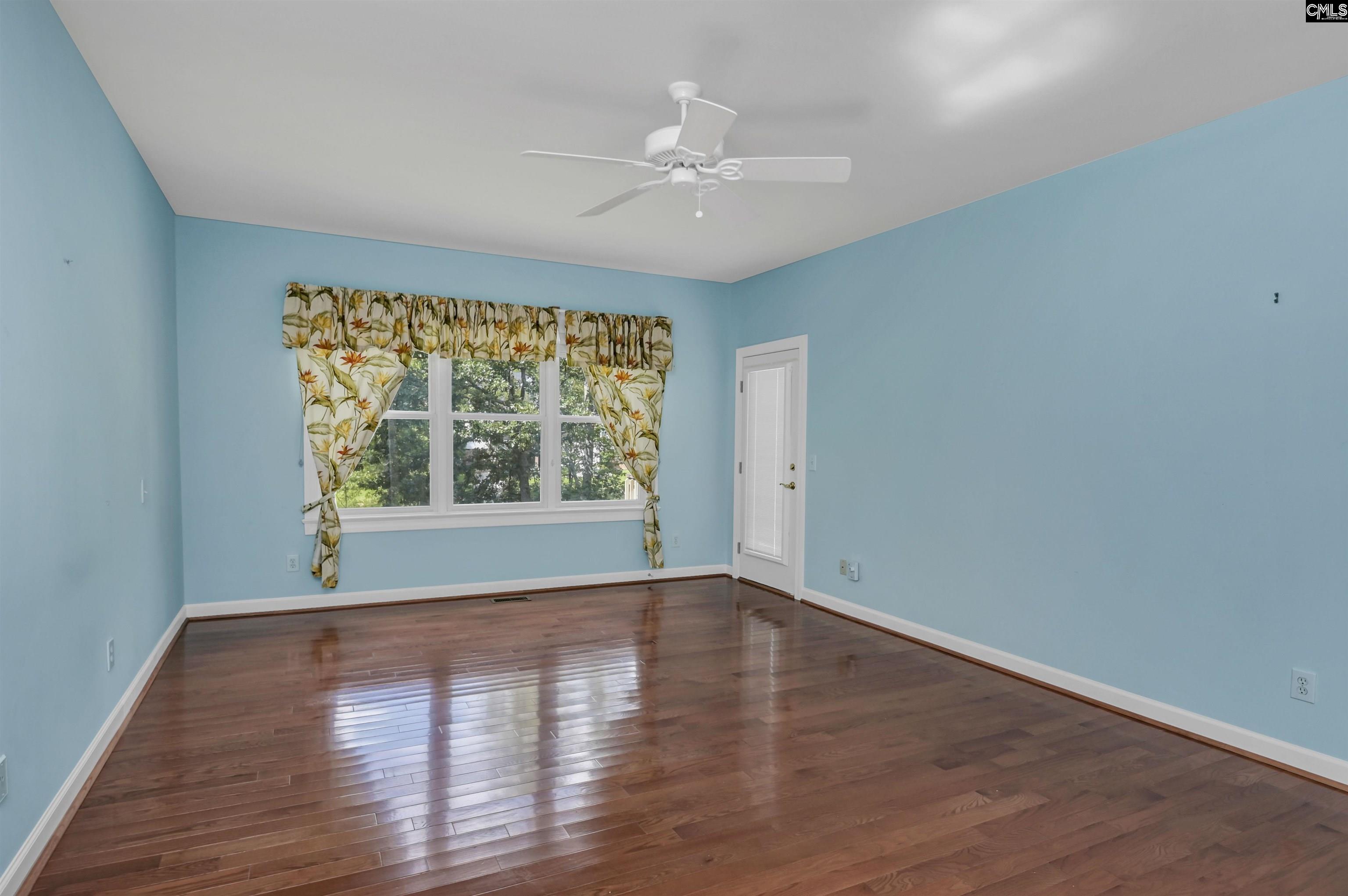 Empty blue room with polished hardwood floors, a ceiling fan, and floral curtains framing the window.