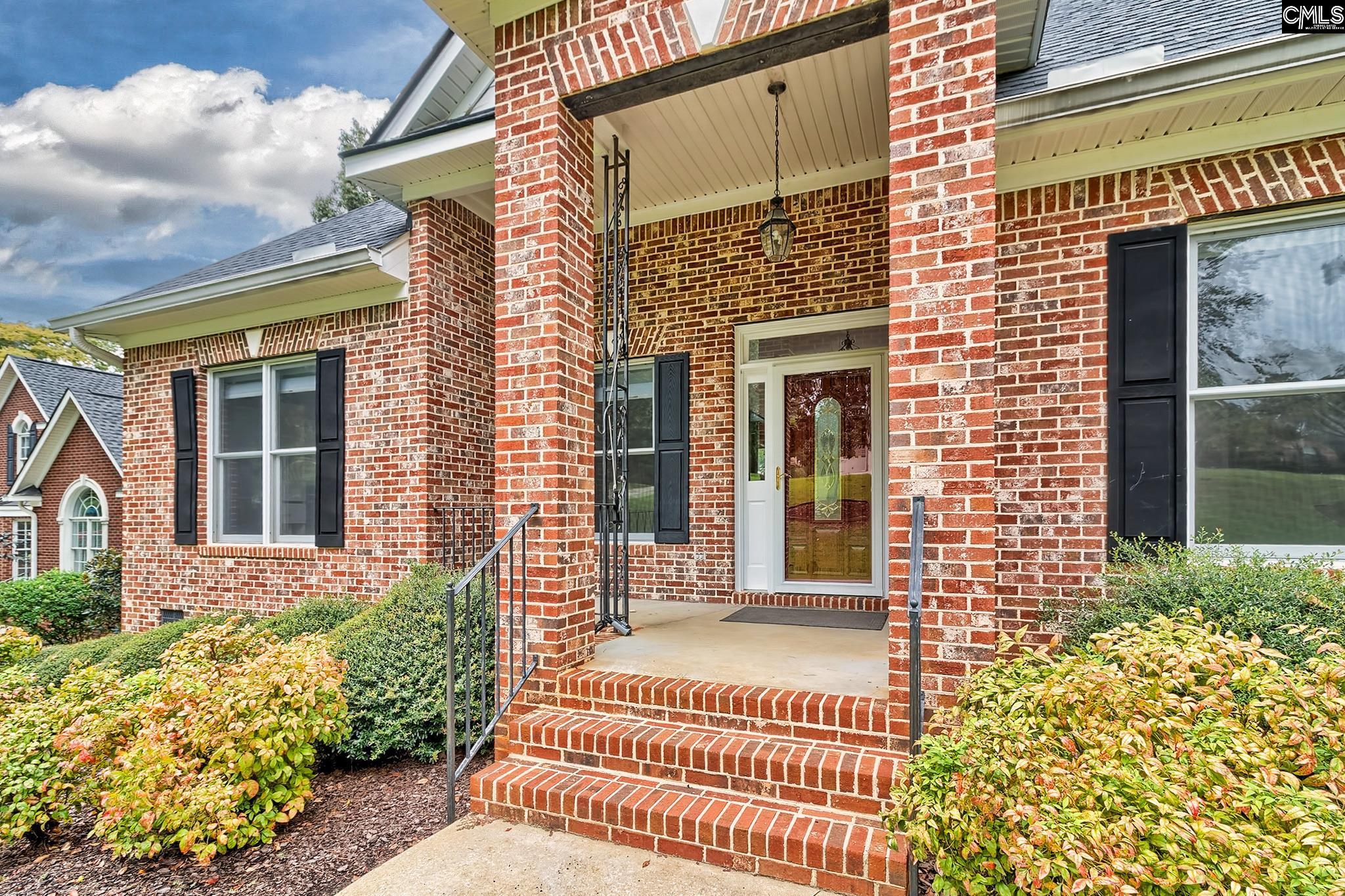 Brick home entrance with a covered porch, glass-paneled front door, brick steps, and landscaped shrubs.
