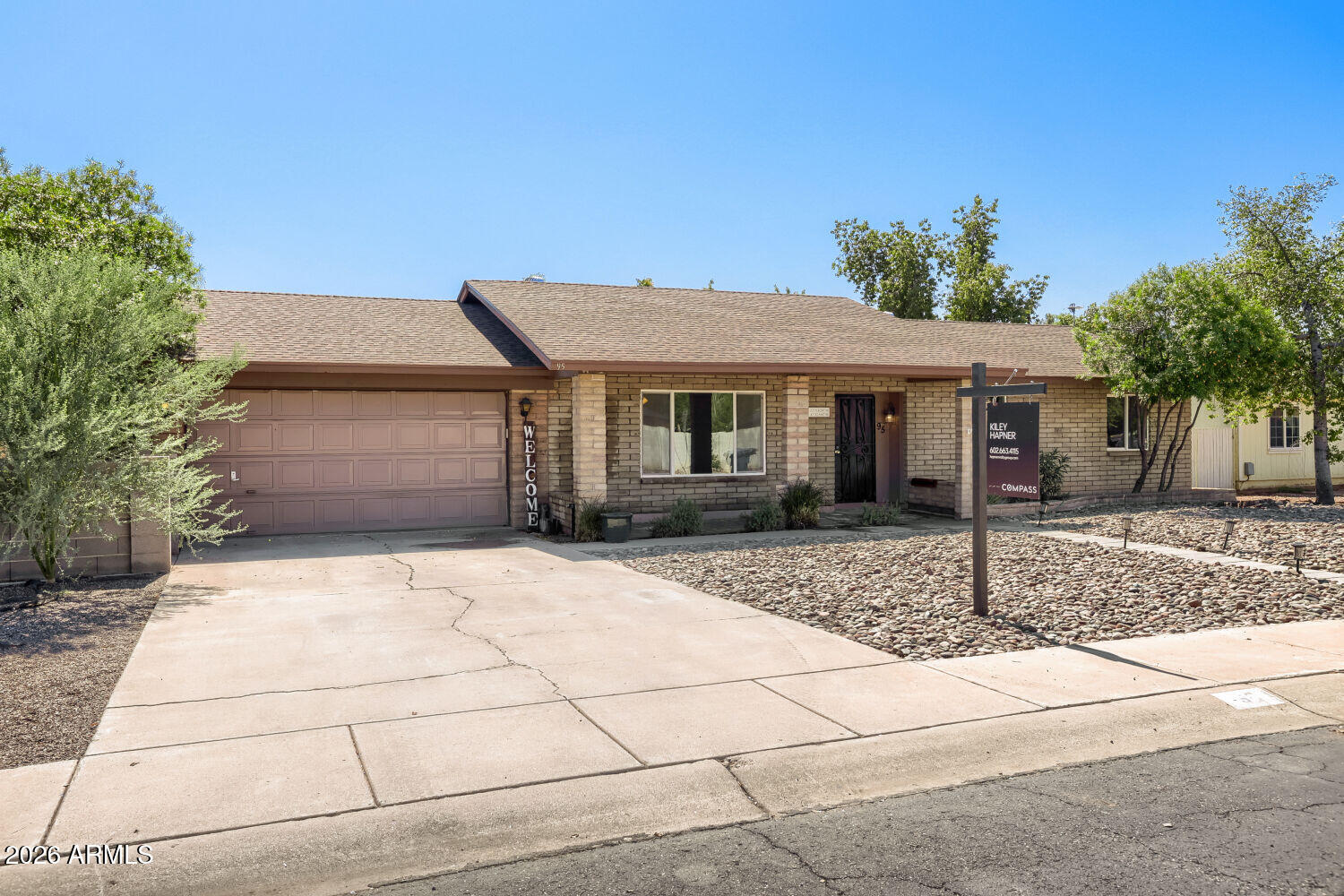 Single-story tan brick home with a two-car garage, desert rock landscaping, and a real estate sign.
