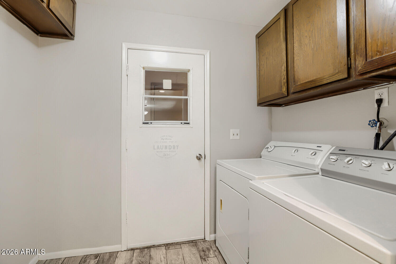 Laundry room with white washer and dryer beneath dark wood cabinets, beside a door with a small window.
