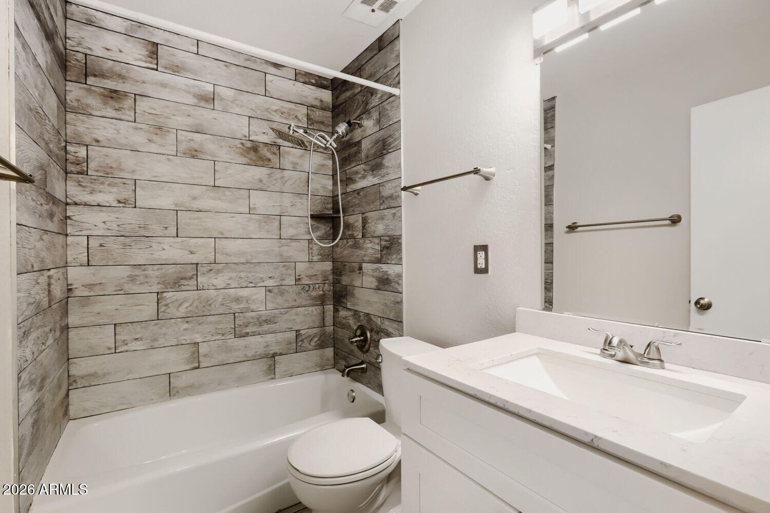 Bathroom with gray wood-look tile around the tub and shower, beside a white vanity and toilet.