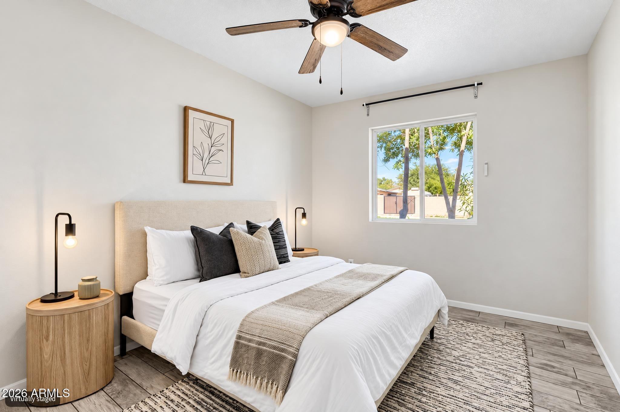 Virtually staged bedroom with a neutral bed, wood bedside tables, ceiling fan, and window overlooking trees.