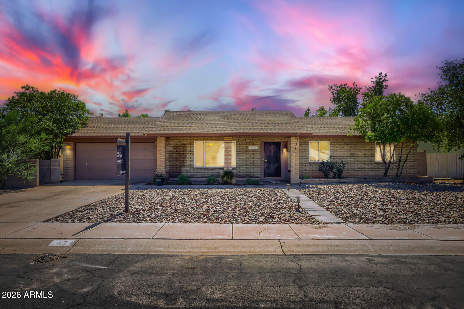 Single-story brick home with a two-car garage and gravel landscaping beneath a colorful sunset sky.