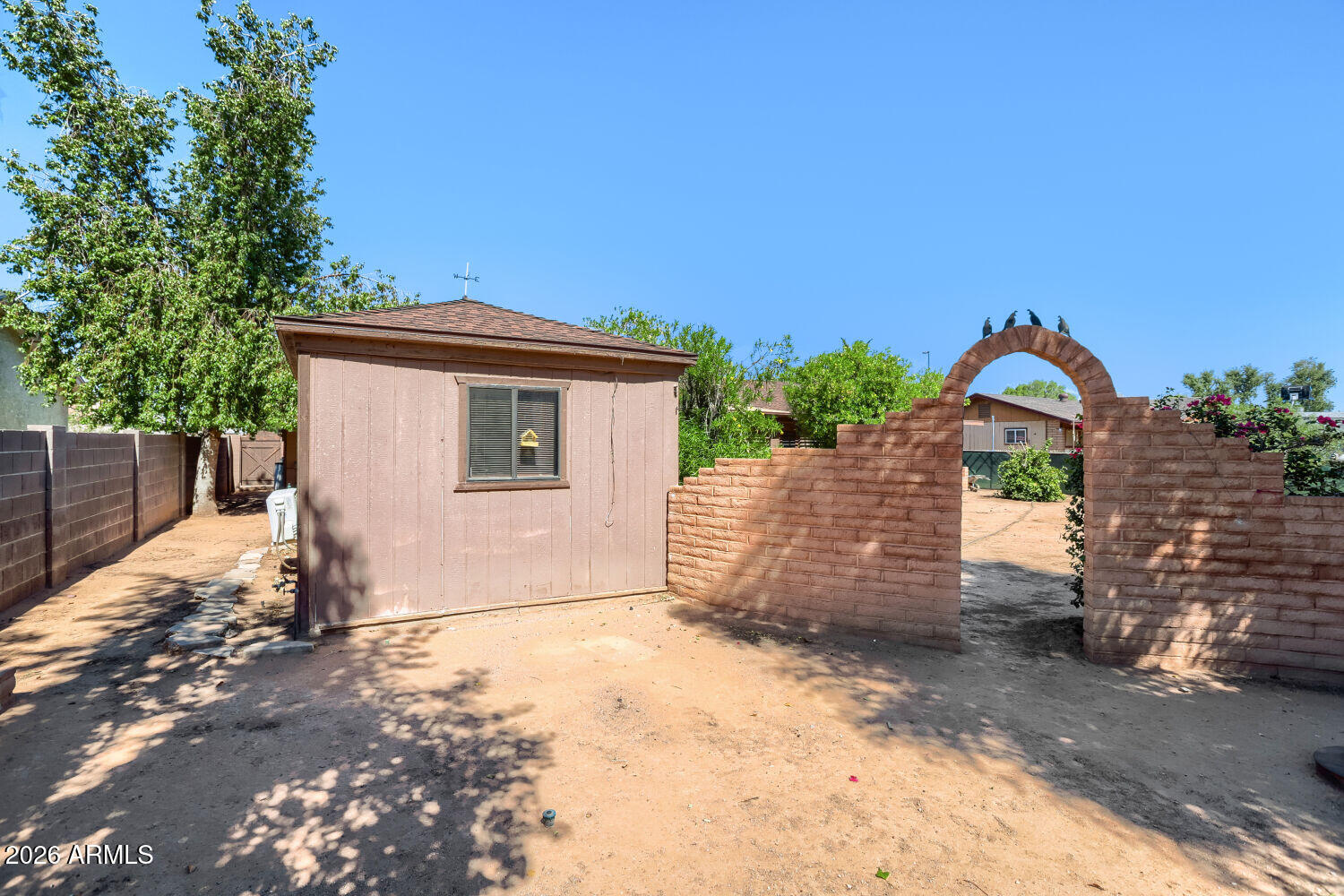 Backyard with a small shed and brick archway opening onto a broad dirt yard.