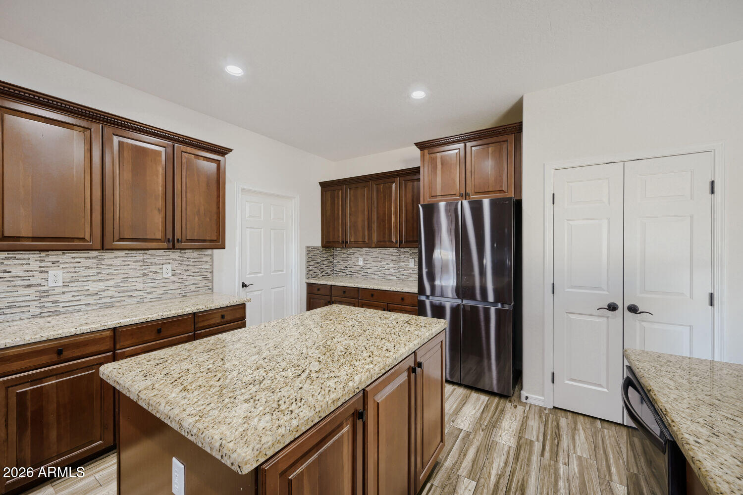 Kitchen with dark wood cabinets, a large granite island, mosaic backsplash, and stainless steel refrigerator.