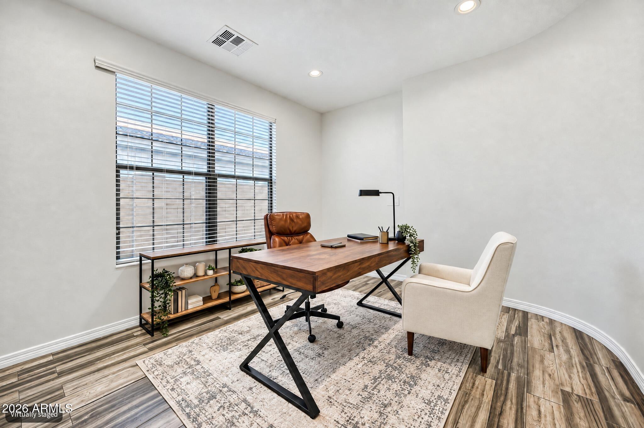 Staged home office with a wooden desk, cream chair, patterned rug, and wide window with blinds.
