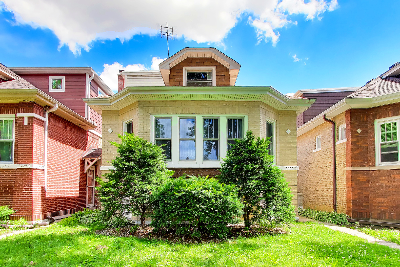 Light brick bungalow with a front dormer, broad windows, and mature shrubs in front.