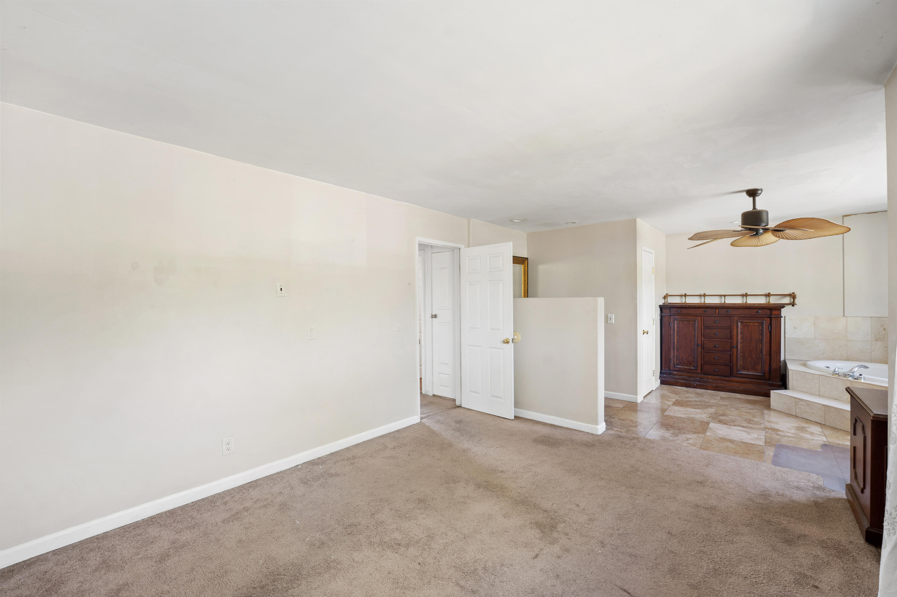 Mostly empty carpeted bedroom opening into a tiled bathroom with a soaking tub and ceiling fan.