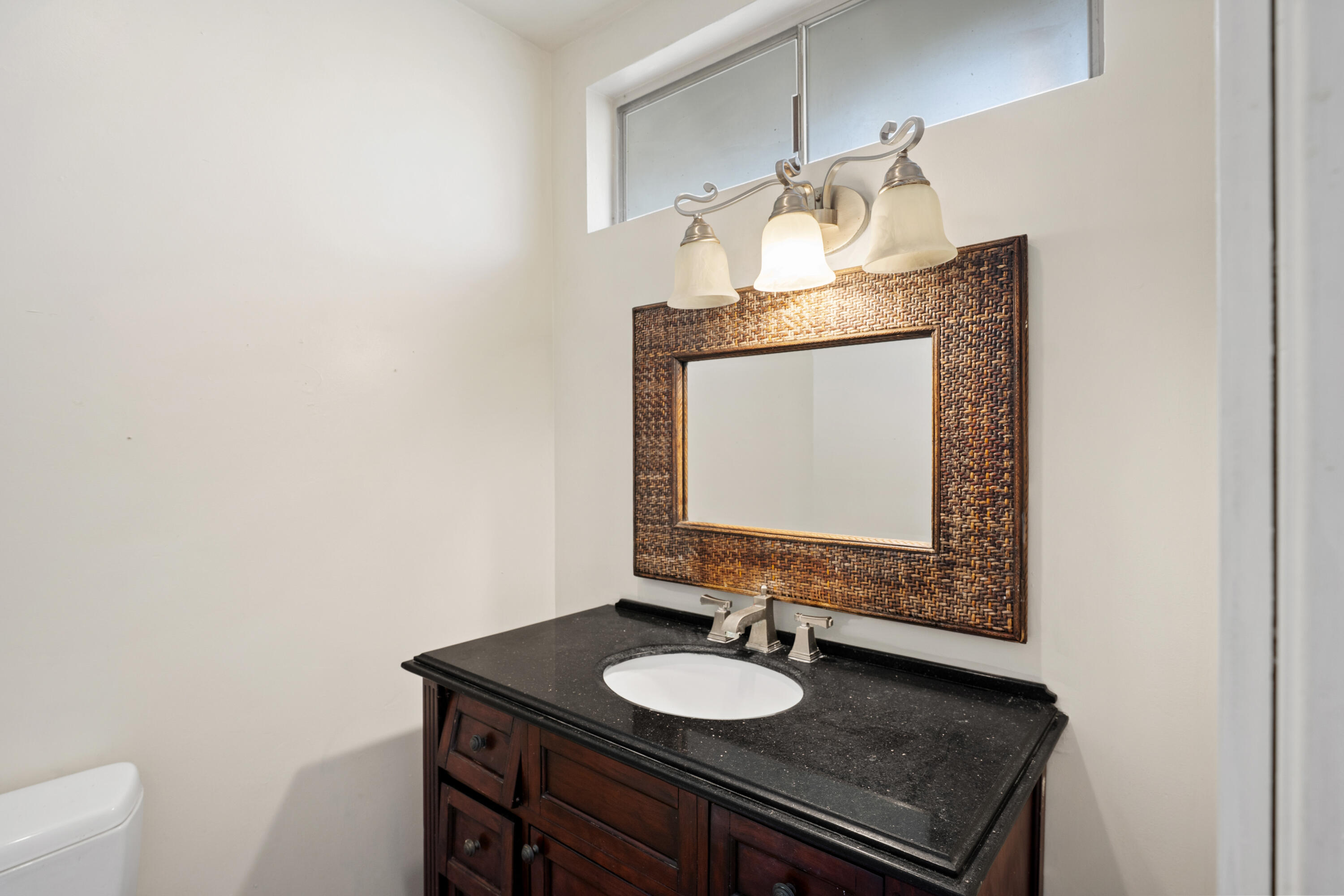 Bathroom vanity with a dark wood cabinet, black countertop, and woven-framed mirror.