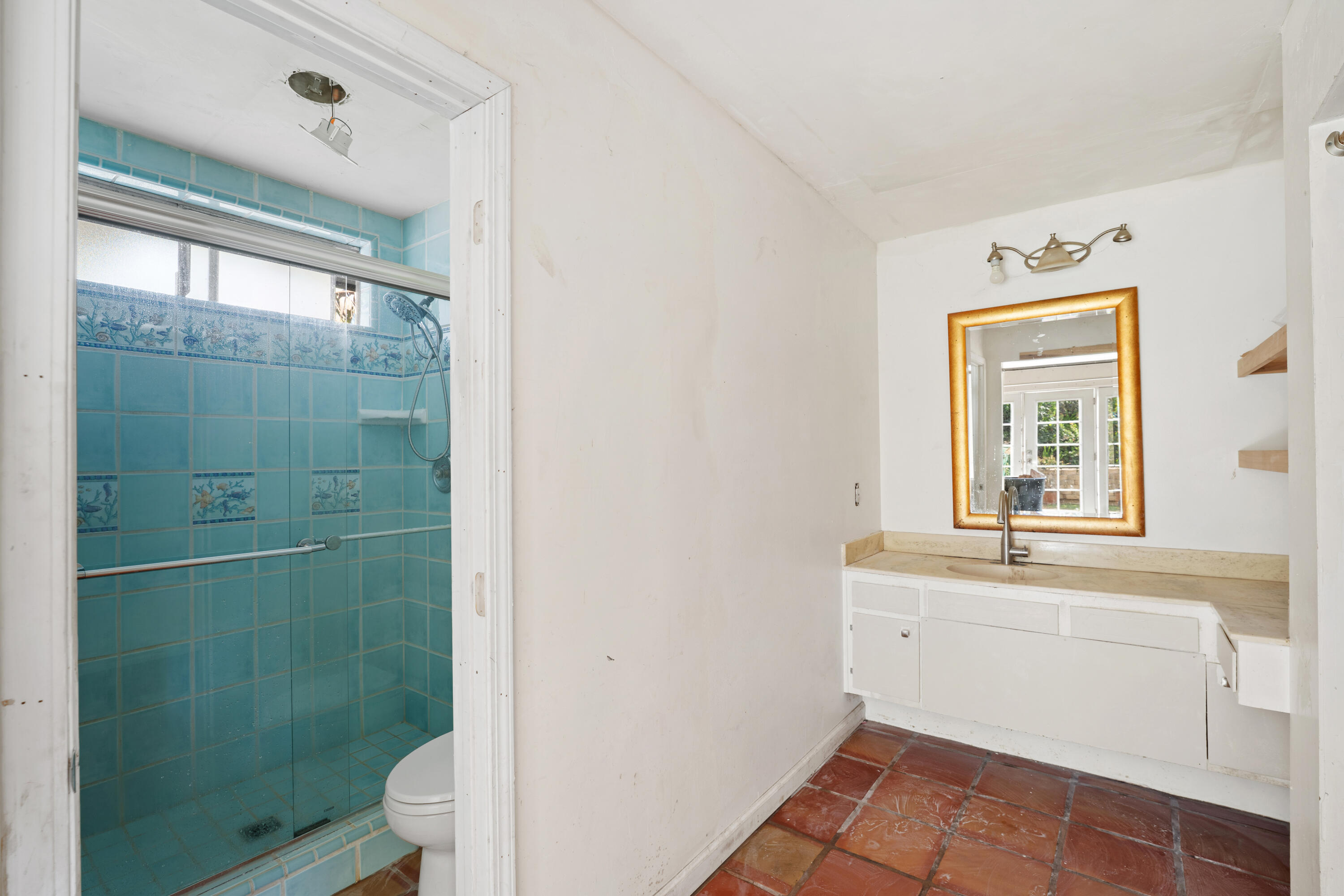 Bathroom with a turquoise tiled shower, white vanity, gold-framed mirror, and terracotta tile floor.