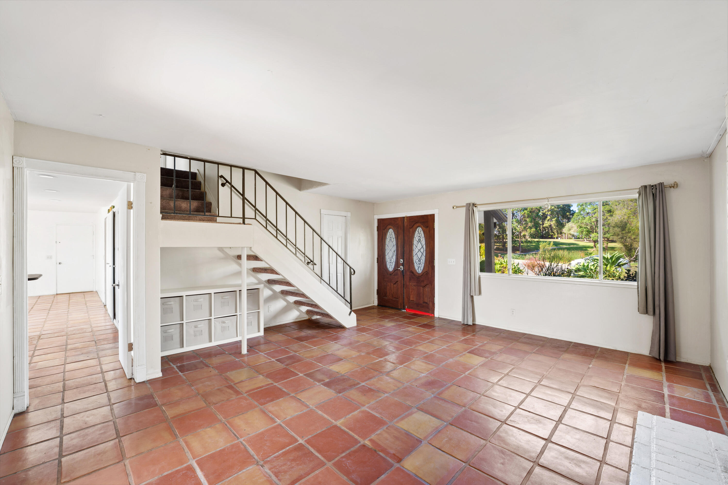 Vacant entry and living area with terracotta tile floors, staircase, carved double doors, and a window overlooking greenery.